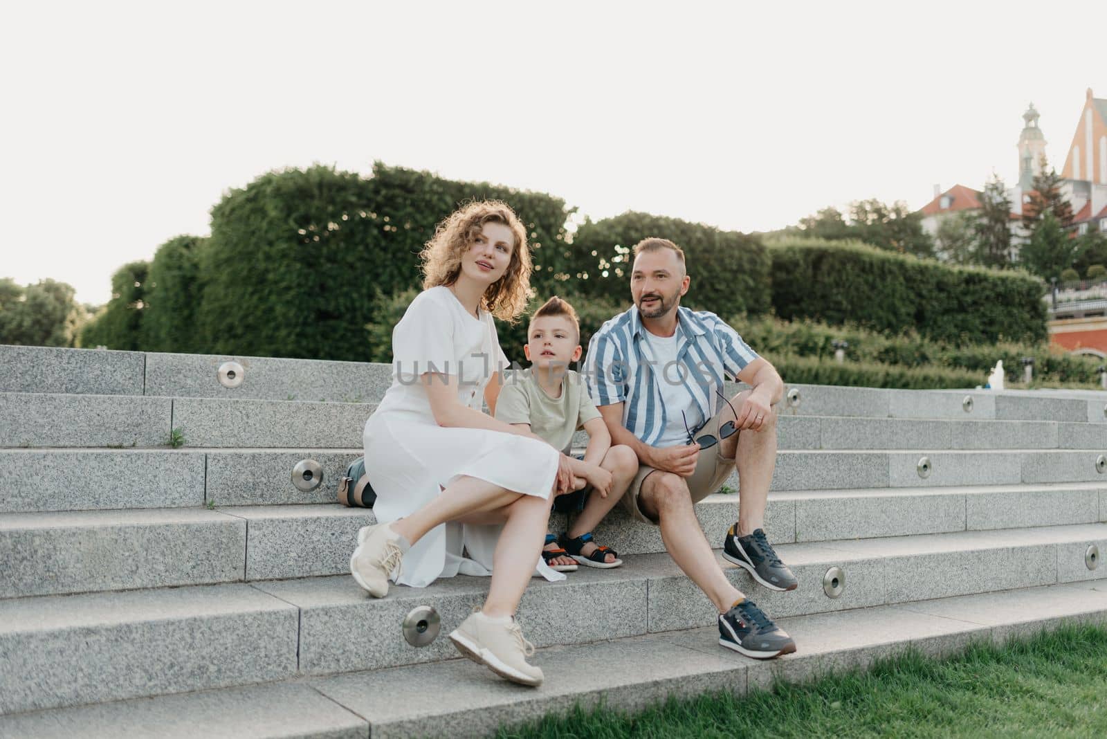 Father, mother, and son are sitting on the steps and staring at the garden of an old European town. Dad is discussing important themes with his smiling family in the park in summer at sunset