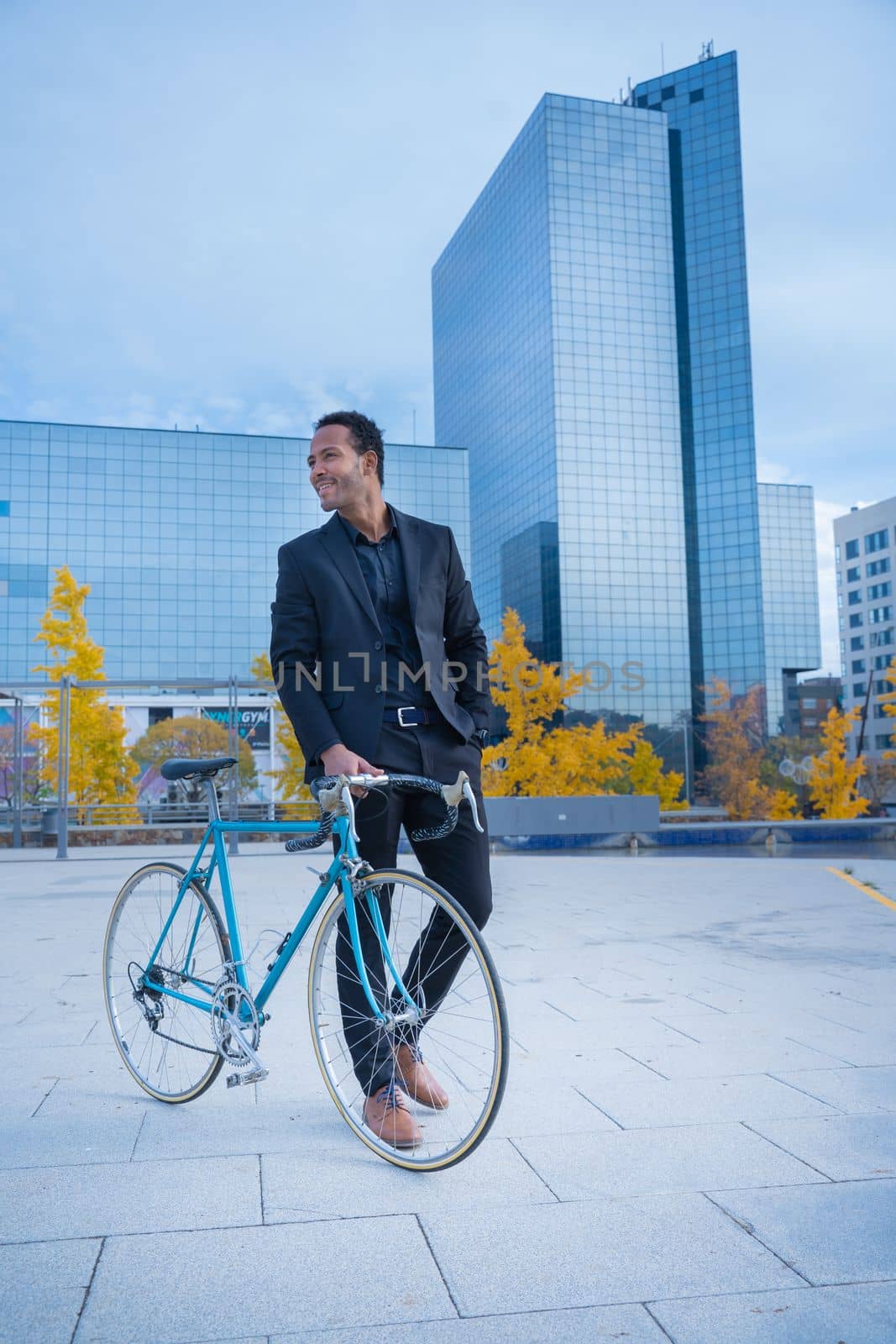 Young smiling stylish African American businessman in suit going to the office by bicycle in front of financial district. Business and transport concept. High quality vertical photo