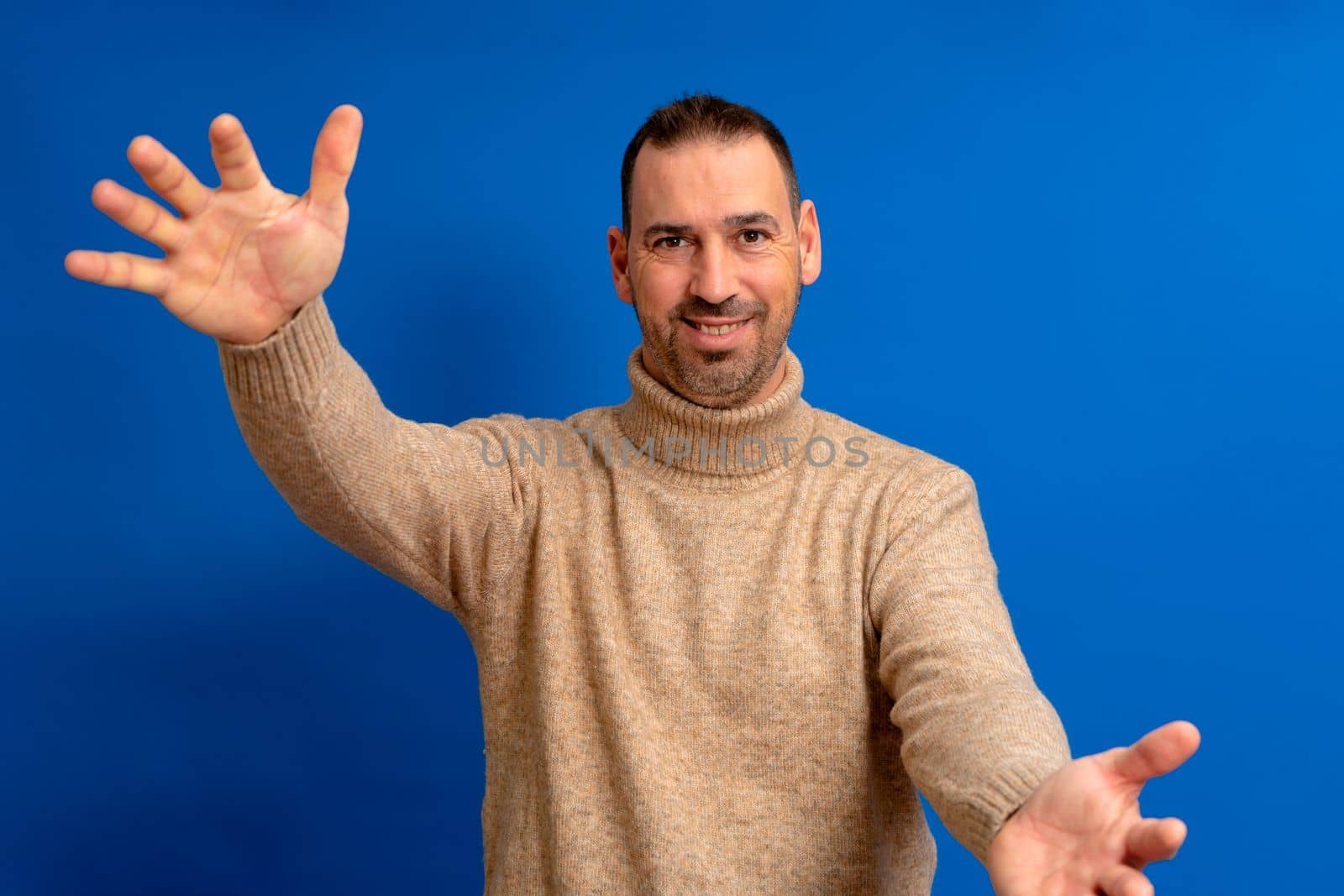 Happy handsome bearded hispanic man standing with arms raised, wants to hug his friend, isolated on blue background... I love you. I miss you body language.