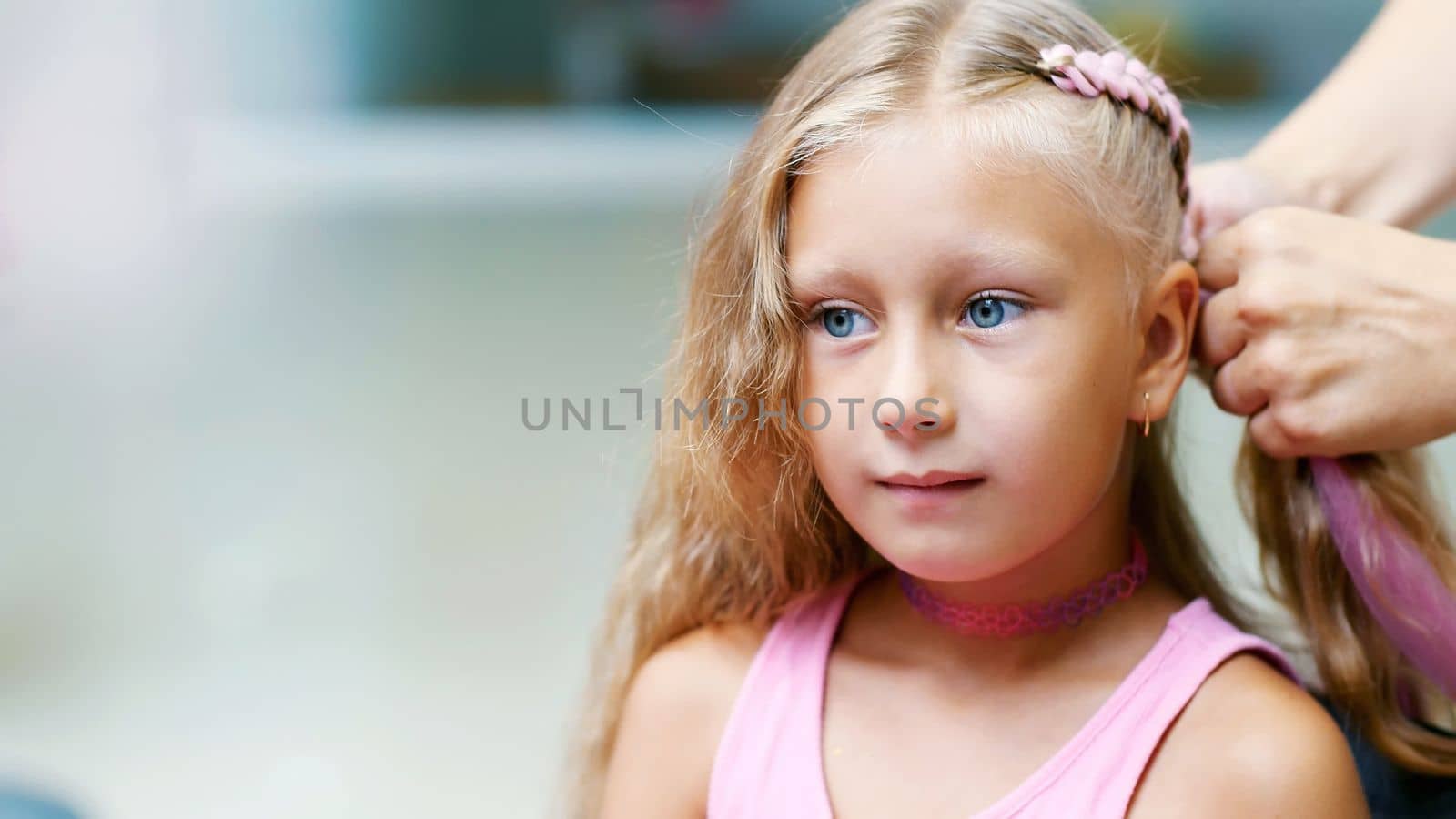 Beautiful blond girl, of seven years old, braided two pigtails, do a hairstyle with pink locks of hair in a beauty salon, a hairdresser's salon, in front of a large mirror. a little princess. High quality photo