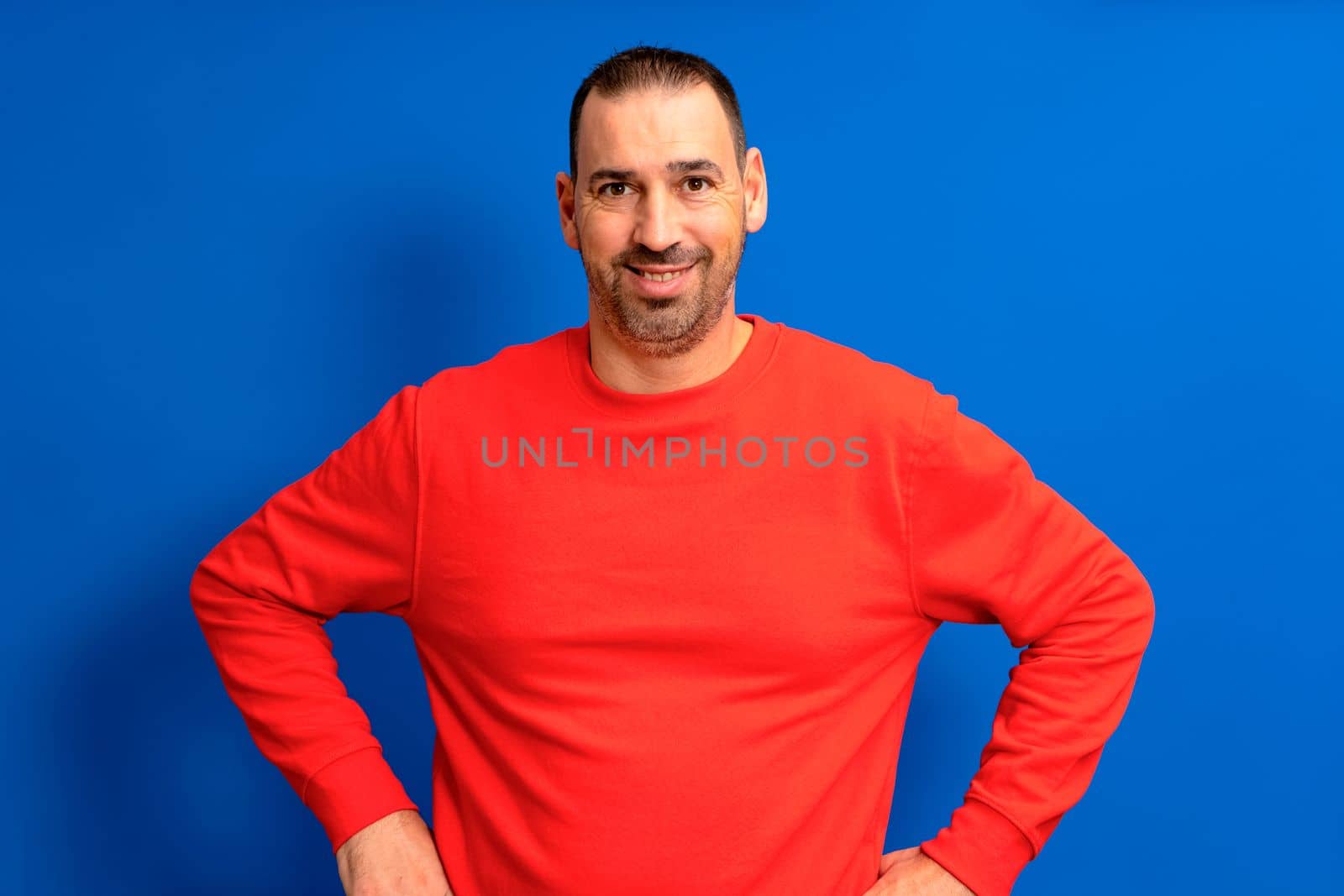 Cheerful bearded man in red sweater posing isolated on a blue background, studio portrait. People lifestyle concept. Mock up copy space. Standing with arms akimbo on waist blinking