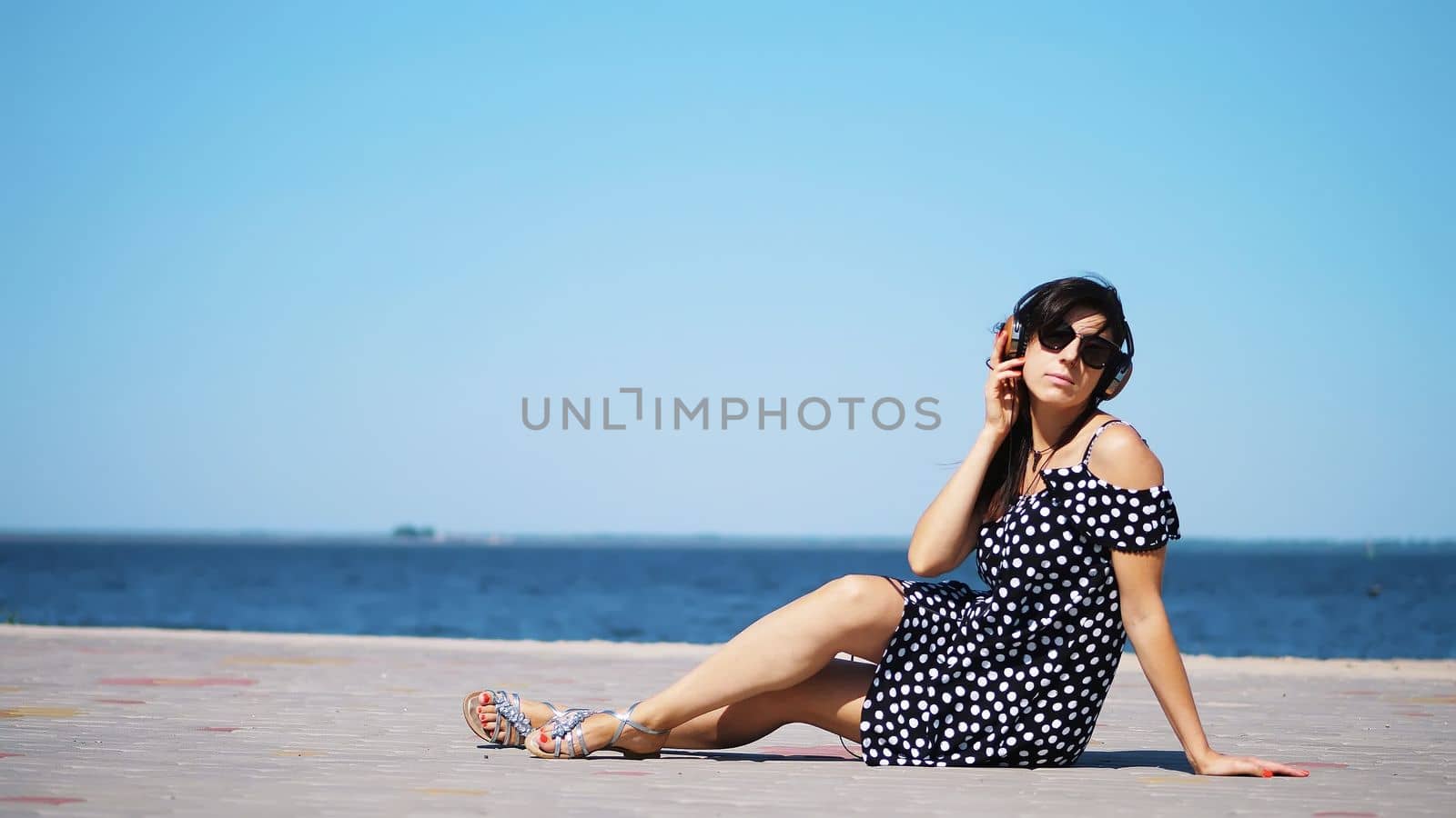 beautiful brunette girl in sun glasses and sundress, with big headphones, listening to music from smartphone, on beach, on hot summer day, near sea. High quality photo