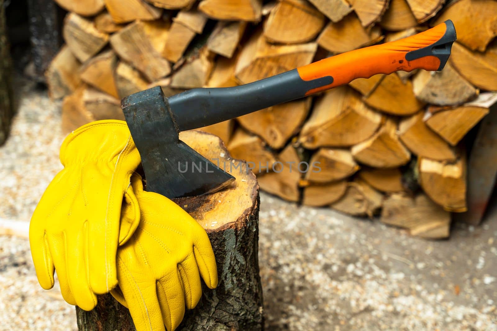 an iron axe stuck in a wooden deck and protective yellow leather gloves against the background of split firewood. Firewood harvesting, preparation for the heating season is a professional tool. An ax is stuck in a wooden deck for chopping firewood