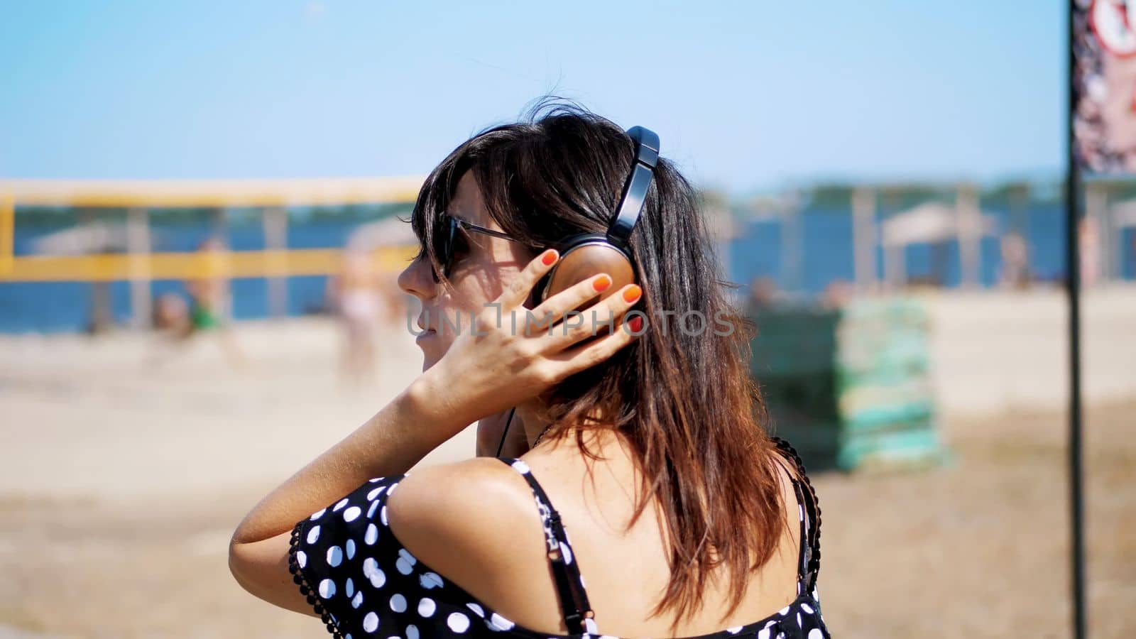 portrait, beautiful girl in sun glasses wearing big black headphones, listening to music from smartphone, on beach, on hot summer day, against background of beach volleyball. High quality photo