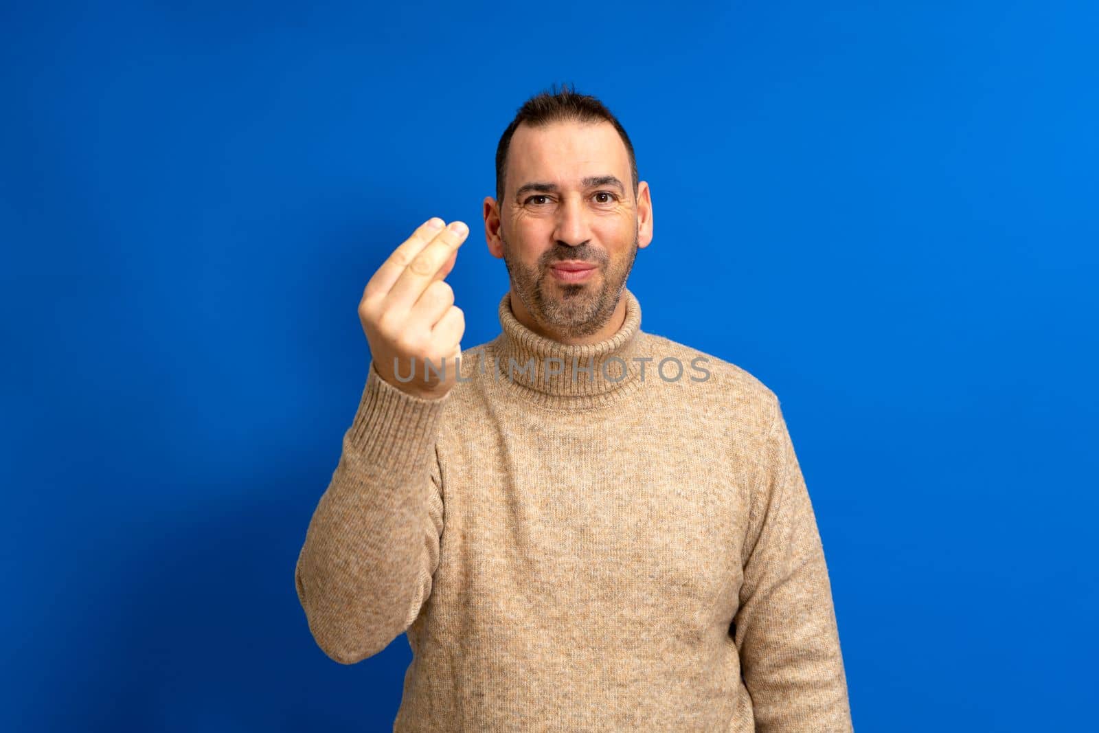 Handsome hispanic man with beard wearing brown turtleneck sweater over blue isolated background doing italian gesture with hand and fingers confident expression