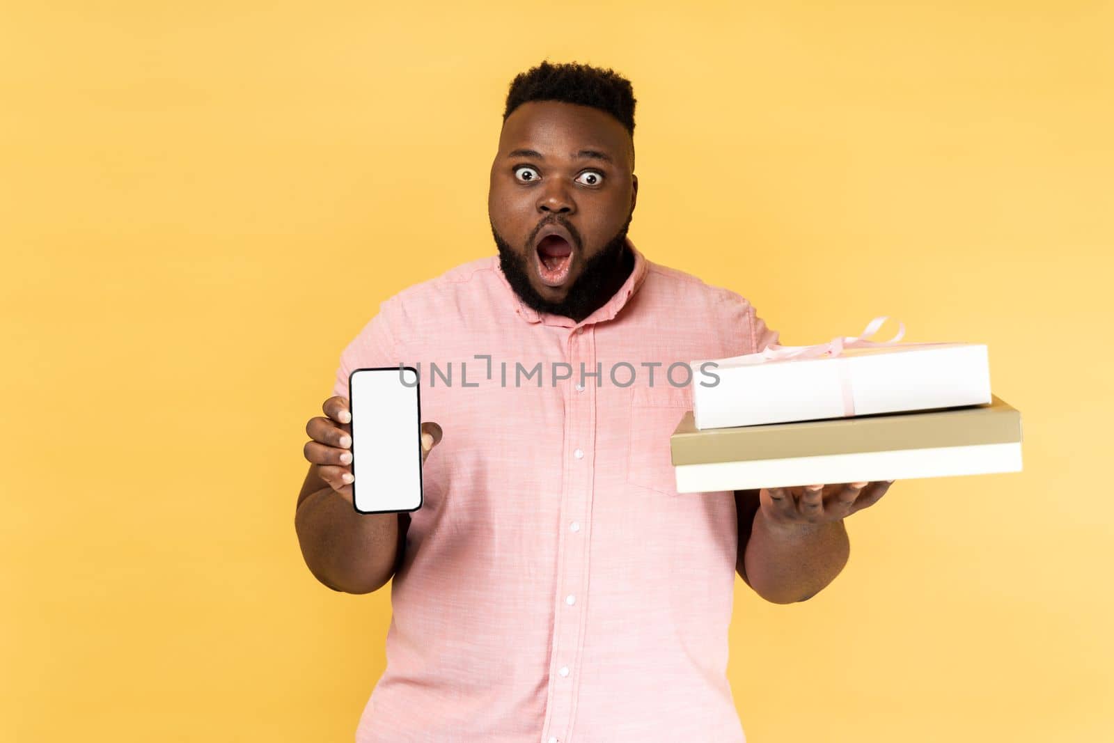 Portrait of shocked astonished man in pink shirt holding present box and smatrphone with blank screen for promotion, looking at camera with big eyes. Indoor studio shot isolated on yellow background.