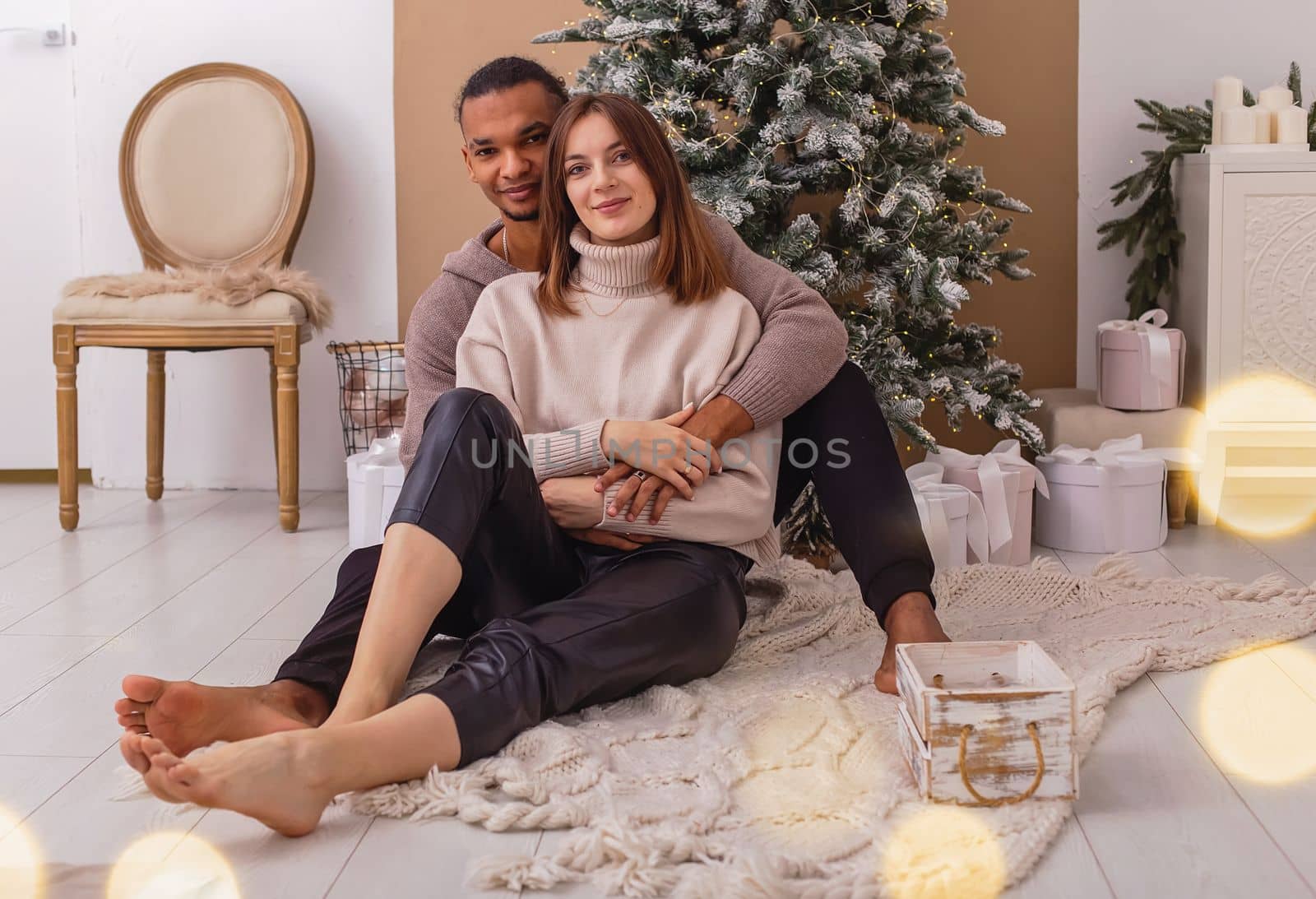 A happy stylish dark-skinned guy in a brown sweatshirt is sitting hugging his girlfriend, near the Christmas tree in the room.