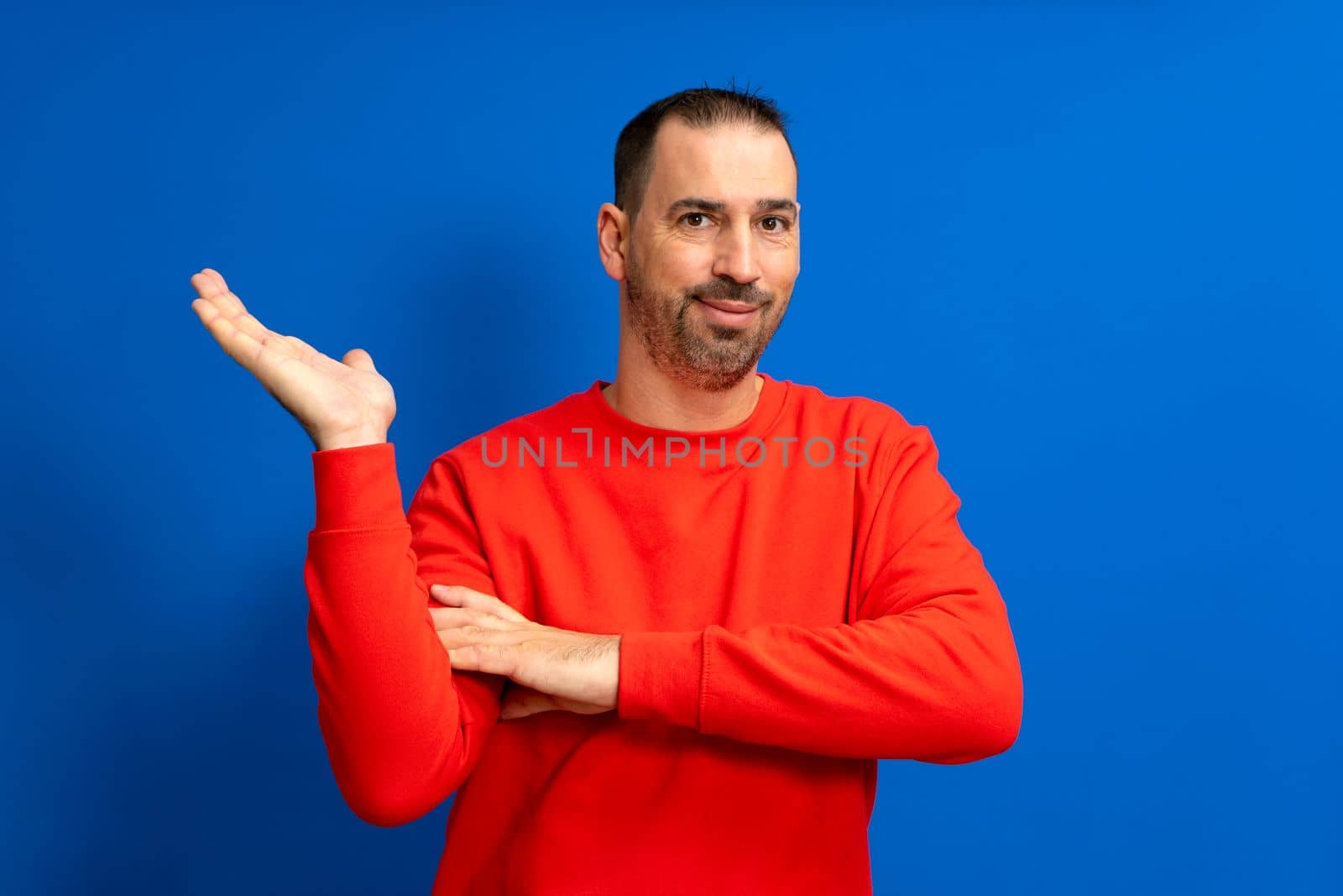 Latino man with beard and red sweater offering something on the palm of his hand, isolated on blue background