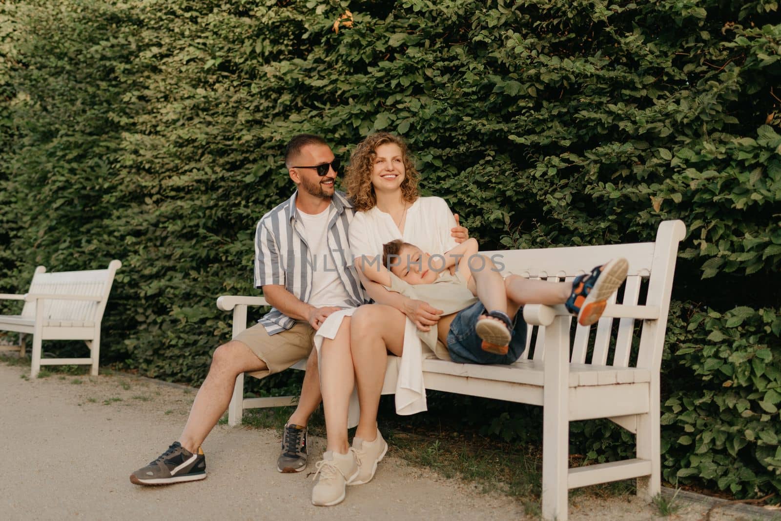 Father, mother, and son are sitting on the bench in the garden of an old European town. Smiling family in the park in summer at sunset