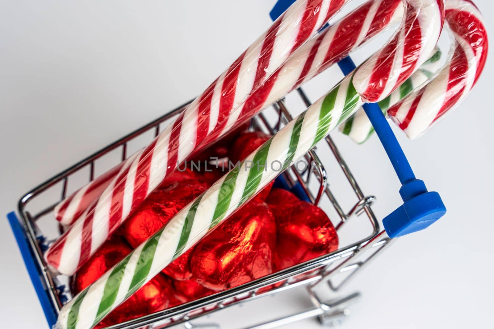 A supermarket trolley loaded with heart-shaped candies and Christmas caramel canes on a white background. Gifts and purchases for the New Year and Christmas. Online trading. A toy shopping cart carries a Christmas candy cane