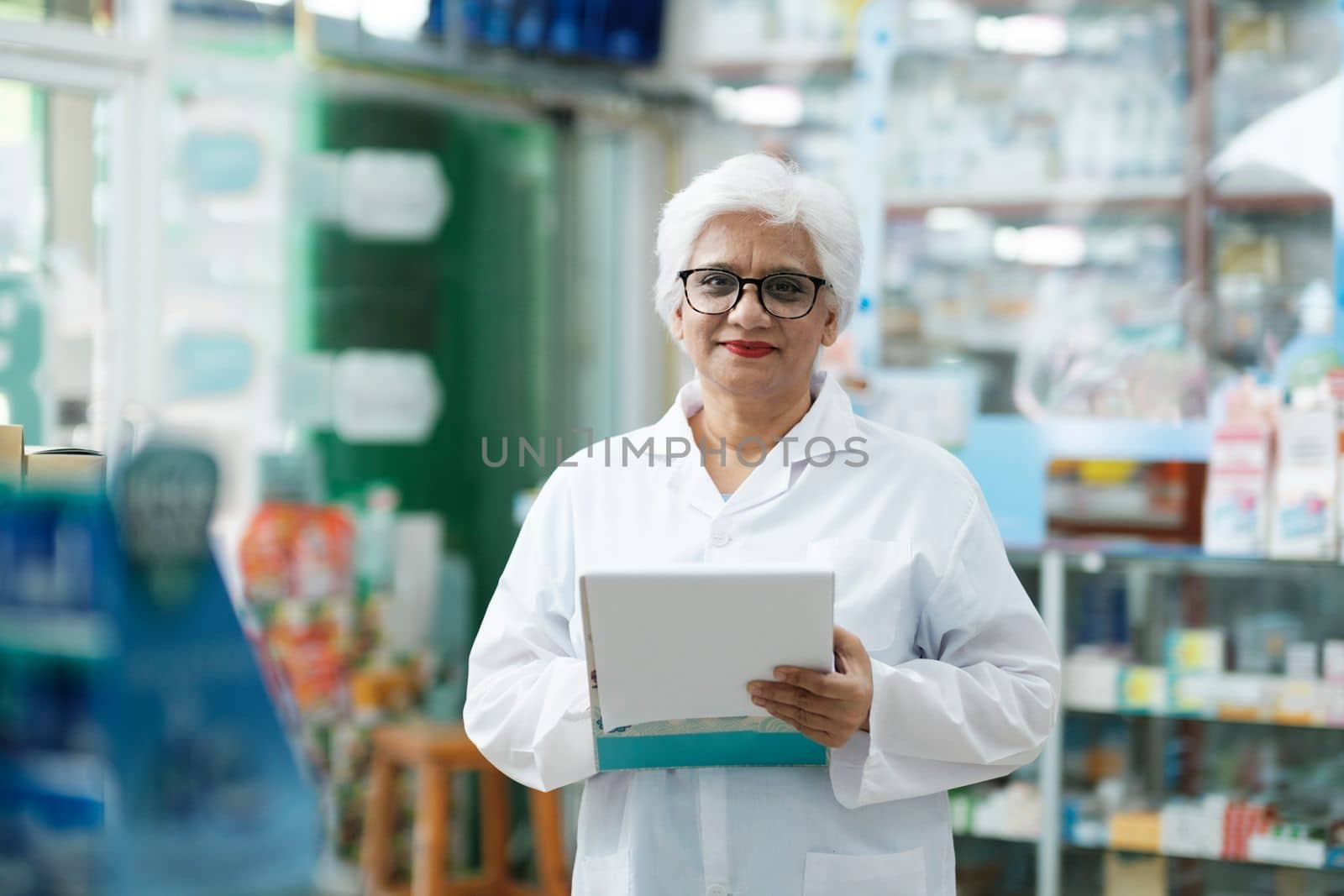 Female smart senior pharmacist in professional white gown looking at camera and checking stock inventory in modern pharmacy, drugstore indoor holding cli. Pharmacy, medicine, and healthcare concept.