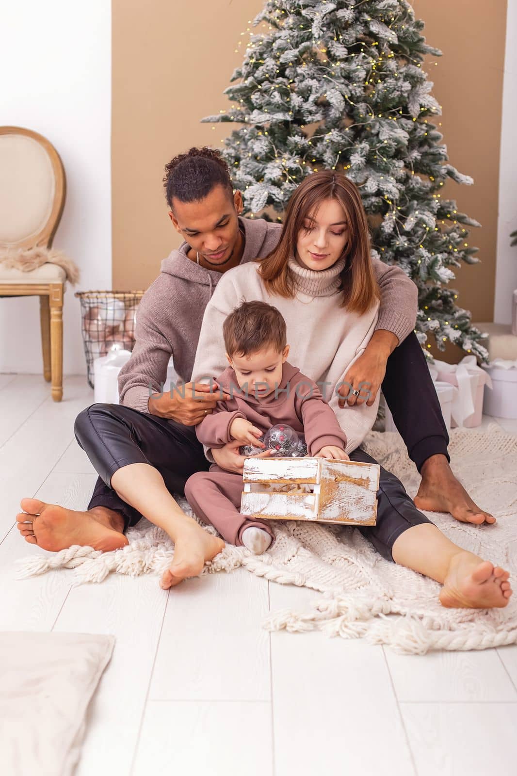 A multi-racial family is sitting on a knitted blanket, holding a wooden box with Christmas toys. by Zakharova