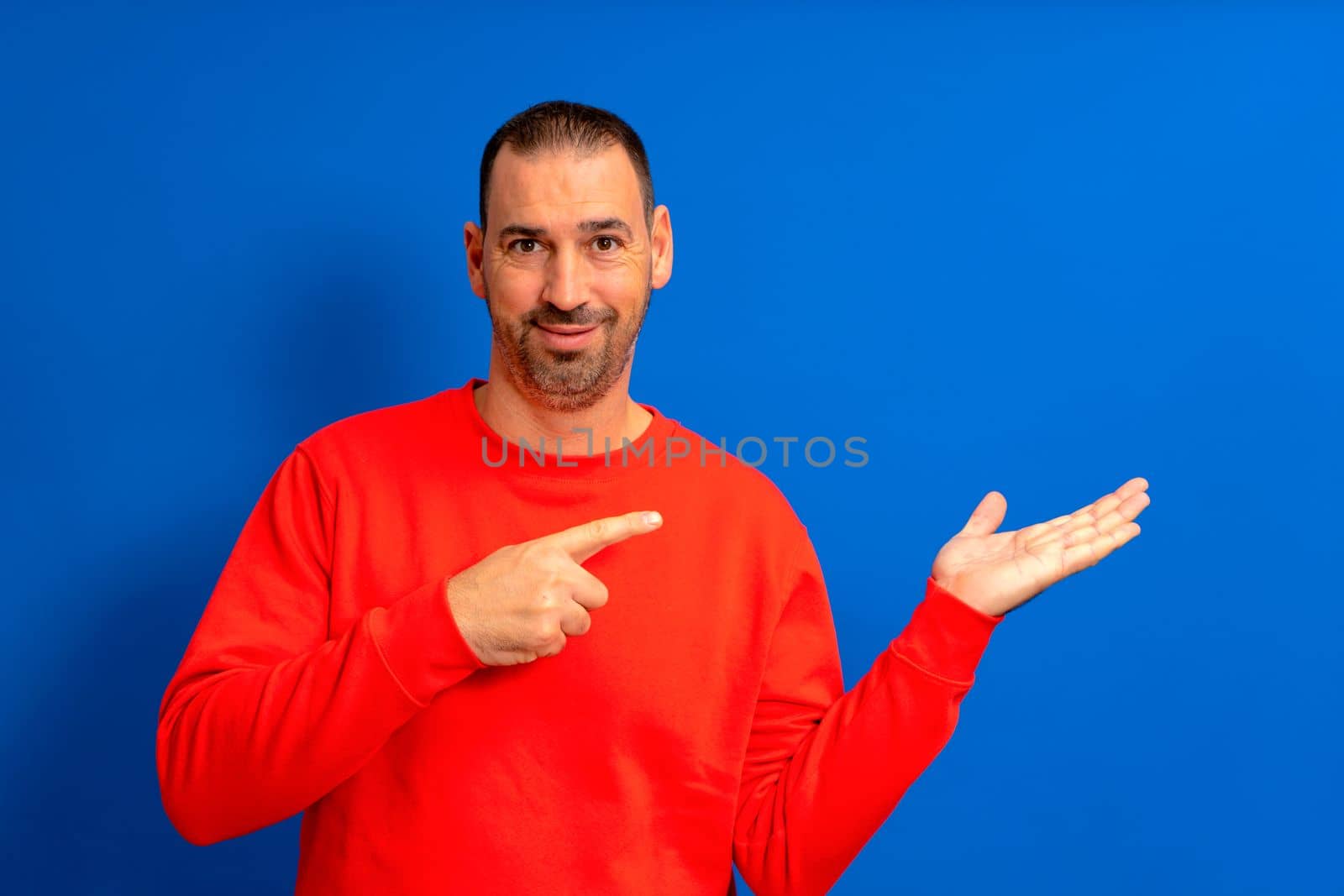 Cheerful bearded man pointing finger holding something on palm. Man in his 40s wearing a red pullover, isolated on a blue background. Sale, advertisement, promotion concept. Close up
