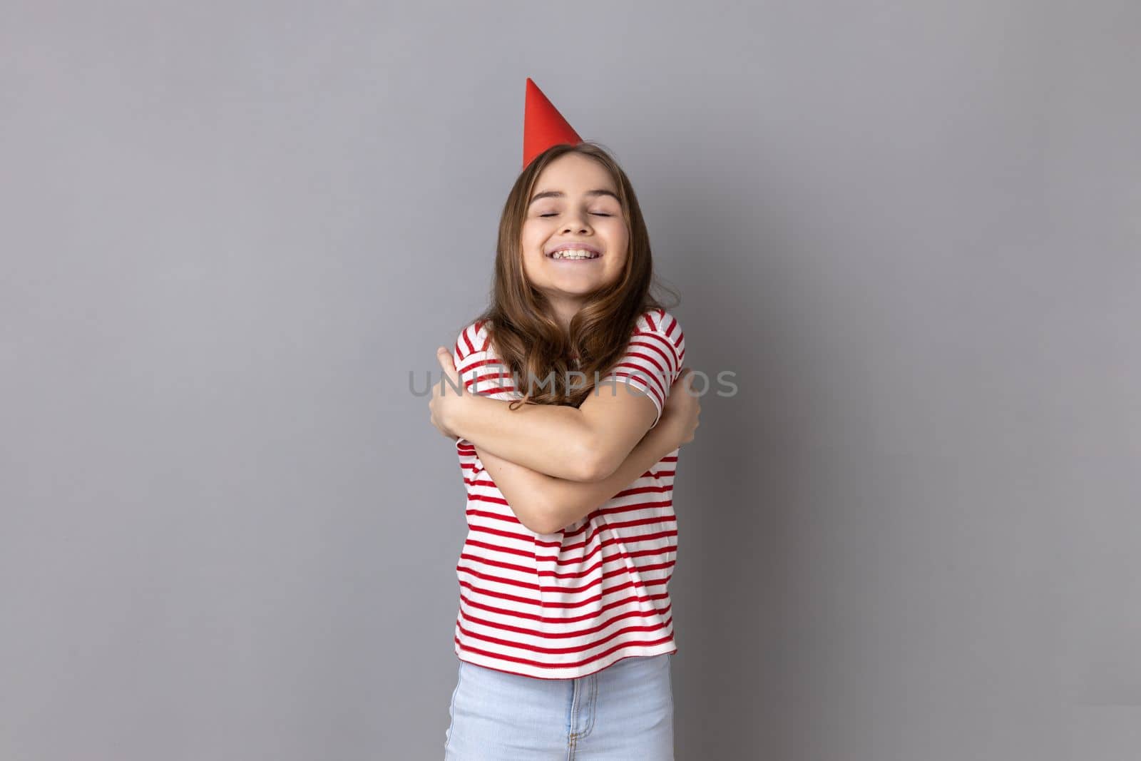 Little girl wearing striped T-shirt and party cone hugging oneself with expression of pleasure and love, celebrating birthday, congratulating herself. Indoor studio shot isolated on gray background.