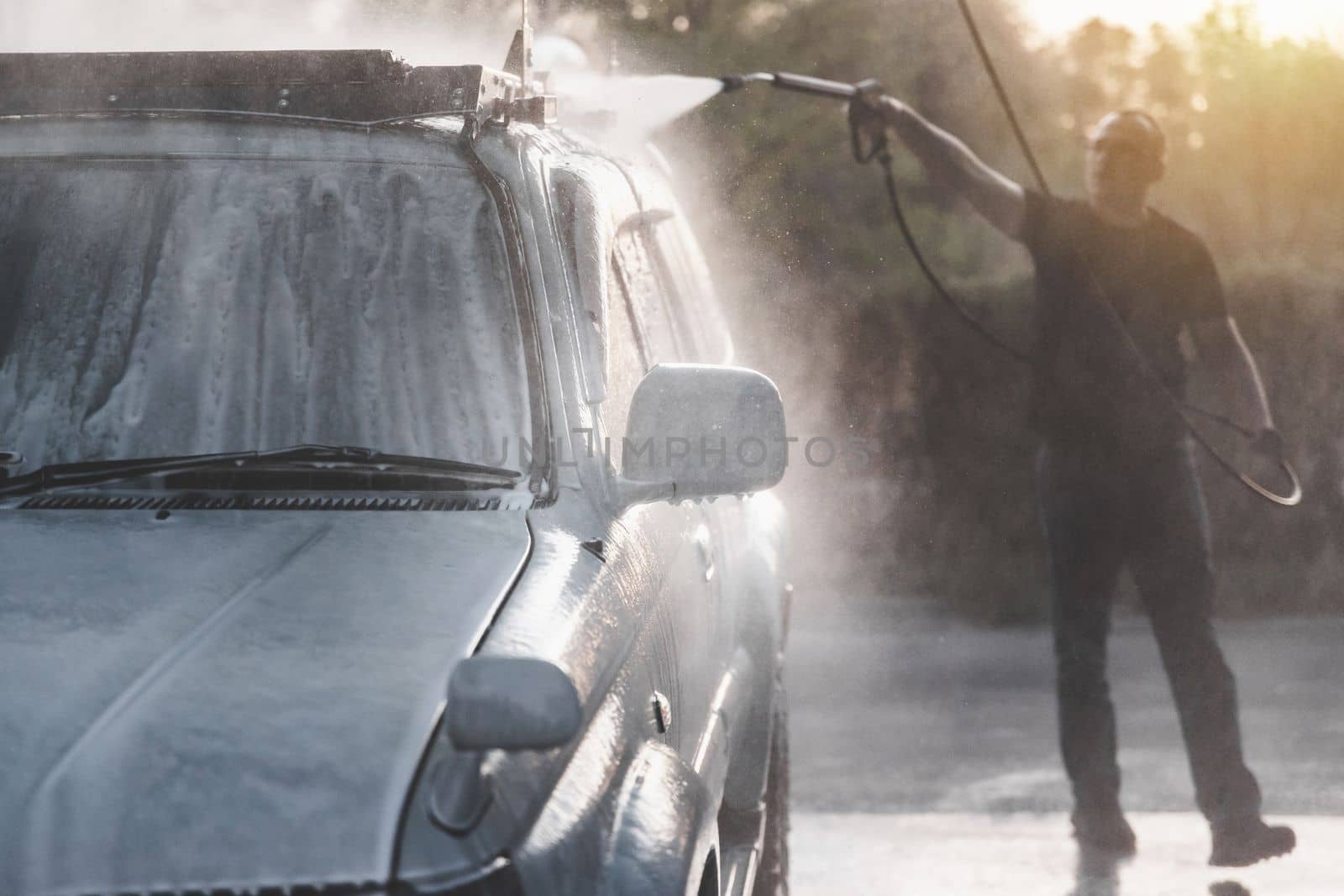 A man with a high pressure hose washes off the foam with water at a self-service car wash.
