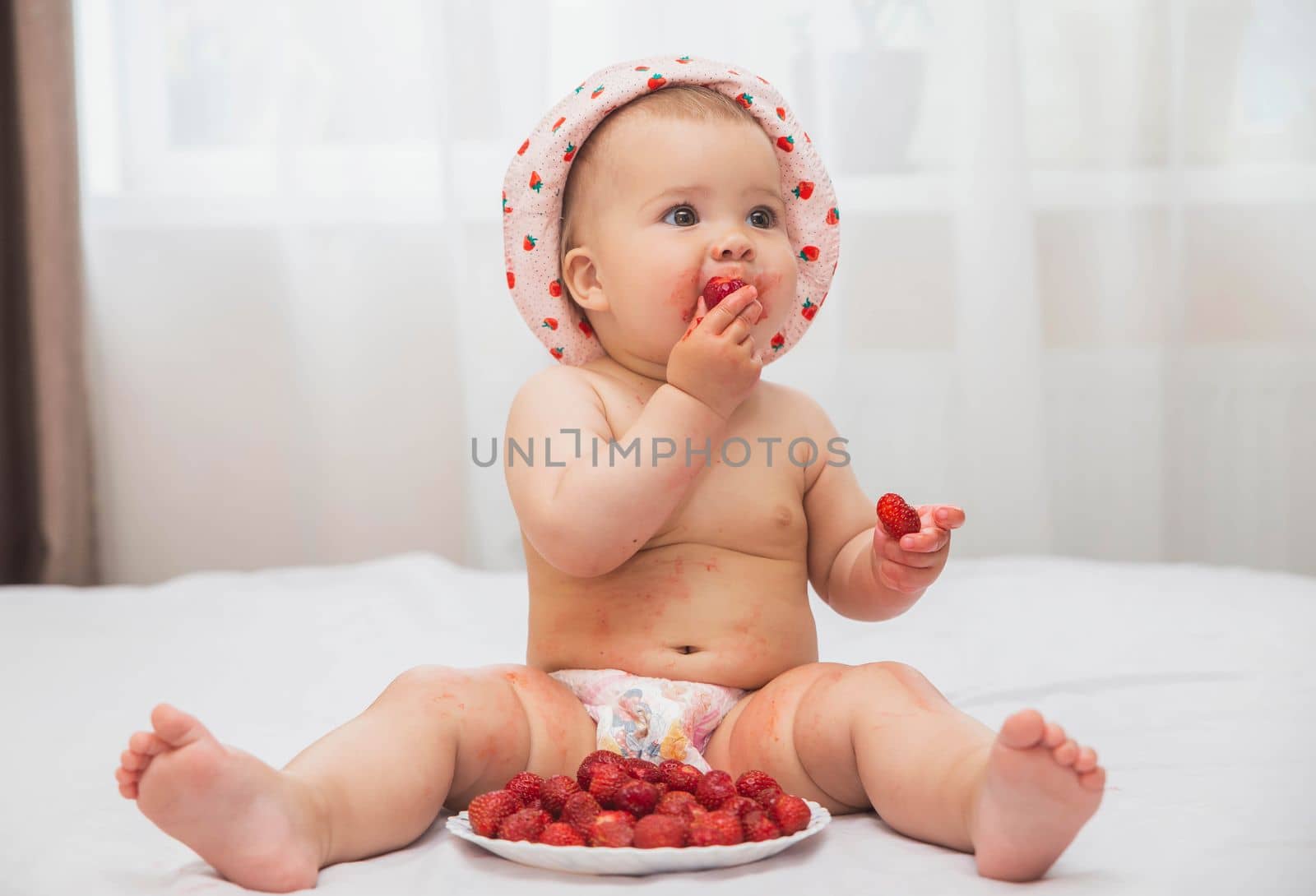 cute stained baby in a hat with painted strawberries eating berries.