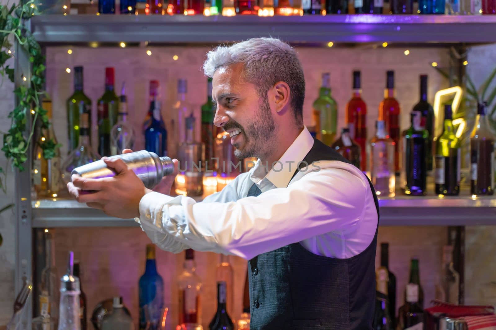Professional bartender in a white shirt and black apron holds a steel shaker in his hands. Blurred bottles on shelves in background. High quality photo