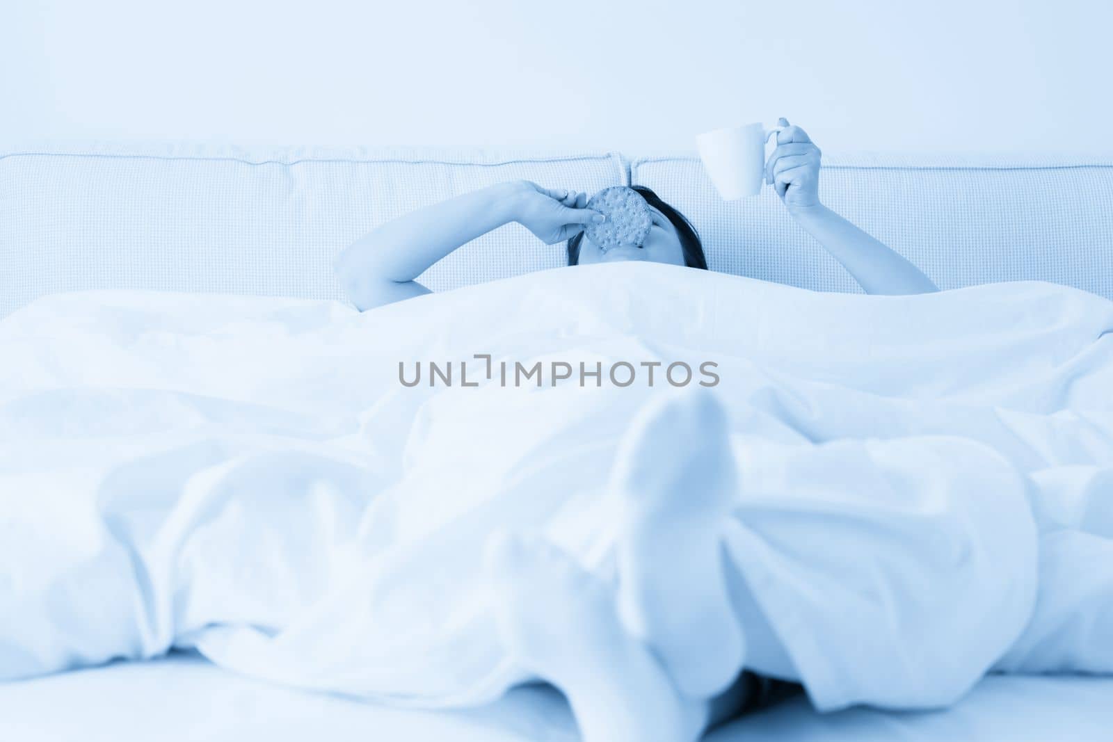Female hand holding cup of coffee from under a blanket in bed. Woman waking up in the morning by Mariakray