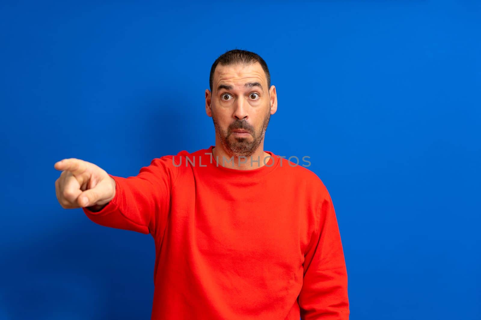 Bearded latino man in a red pullover pointing finger at the camera isolated over blue background