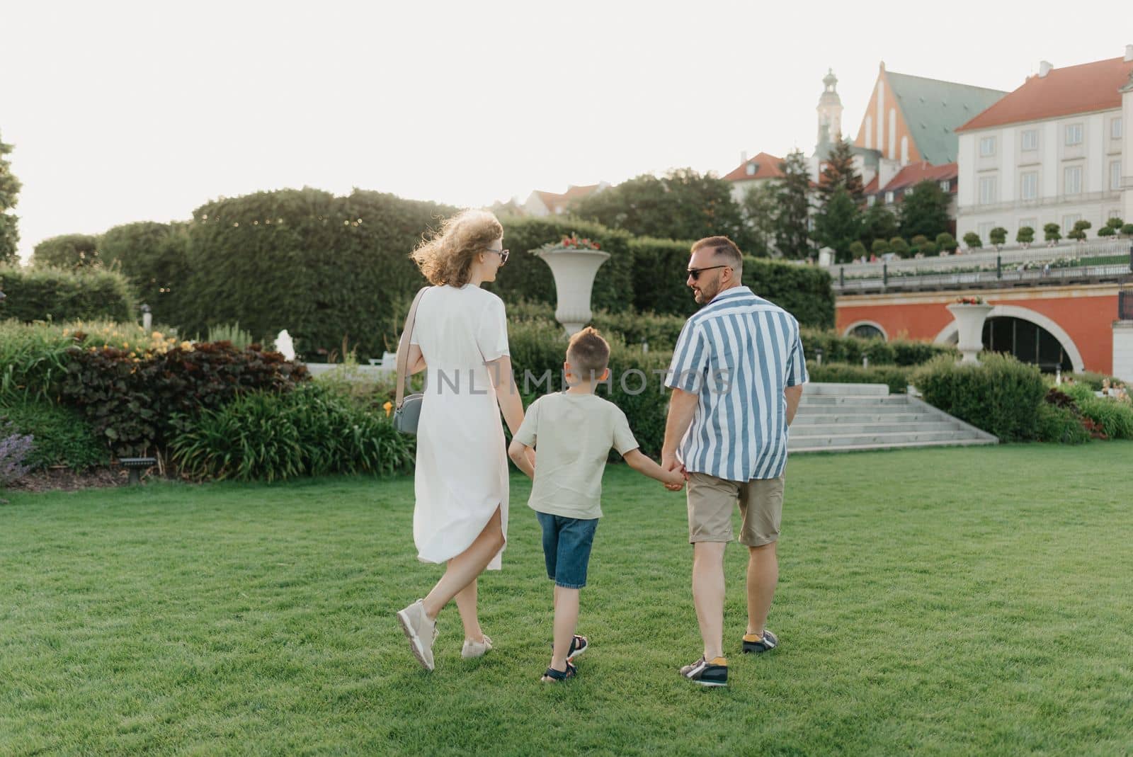 A photo from the back of a family is running in the garden of the palace in an old European town. A happy father, mother, and son are holding hands and having fun at sunset.