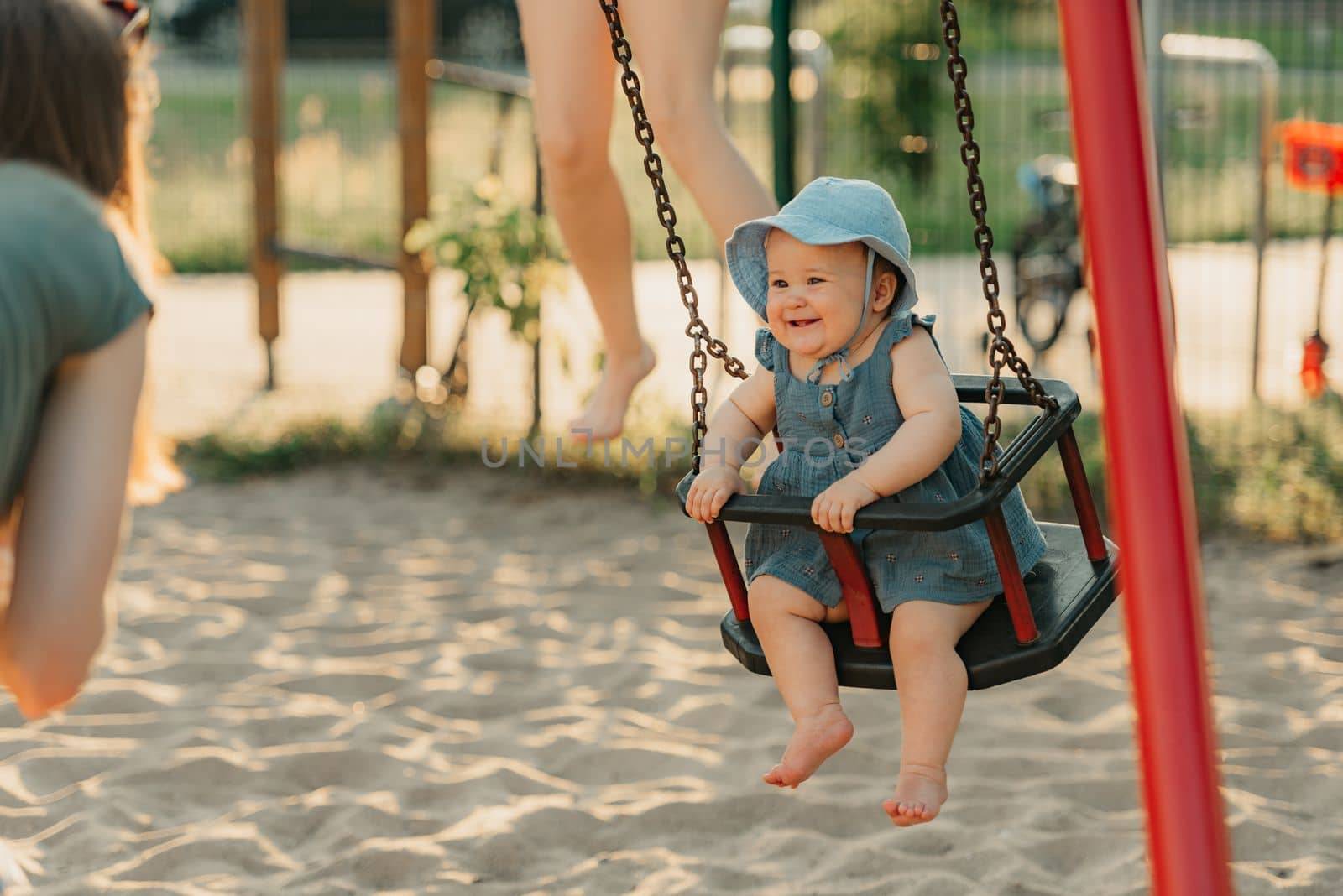 Toddler baby girl on a swing on the warm summer evening. Mother is swinging her young daughter on a sunny playground.