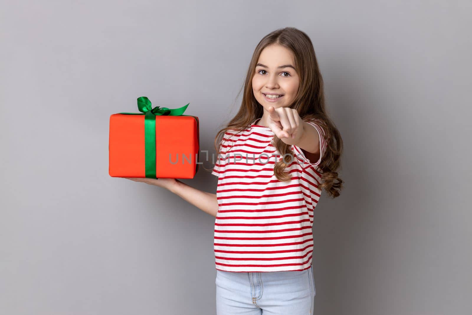 Hey, you are winner. Portrait of delighted little girl wearing striped T-shirt holding present box and pointing finger to camera with excited expression. Indoor studio shot isolated on gray background