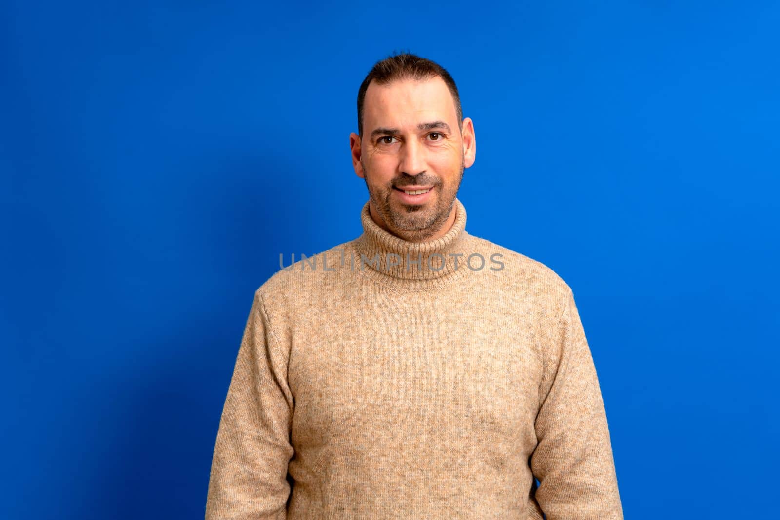 Isolated photo of handsome man with beard, mustache and trendy hairstyle, wears brown turtleneck sweater, has joyful expression while listening to his interlocutor, poses in studio blue background