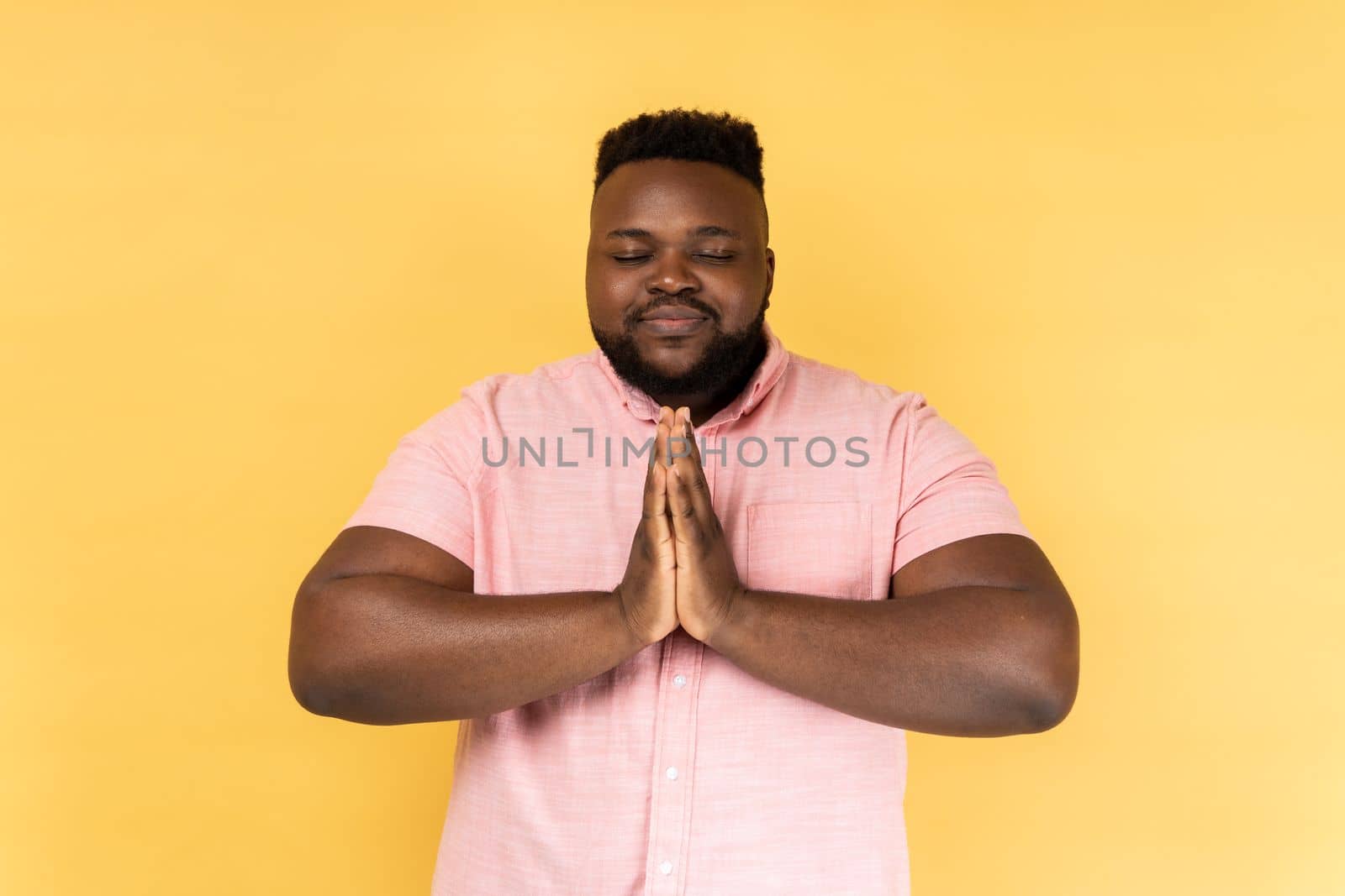 Portrait of man wearing pink shirt keeps hands in yoga gesture, has calm facial expression, keeping palms pressed together and closed eyes. Indoor studio shot isolated on yellow background.