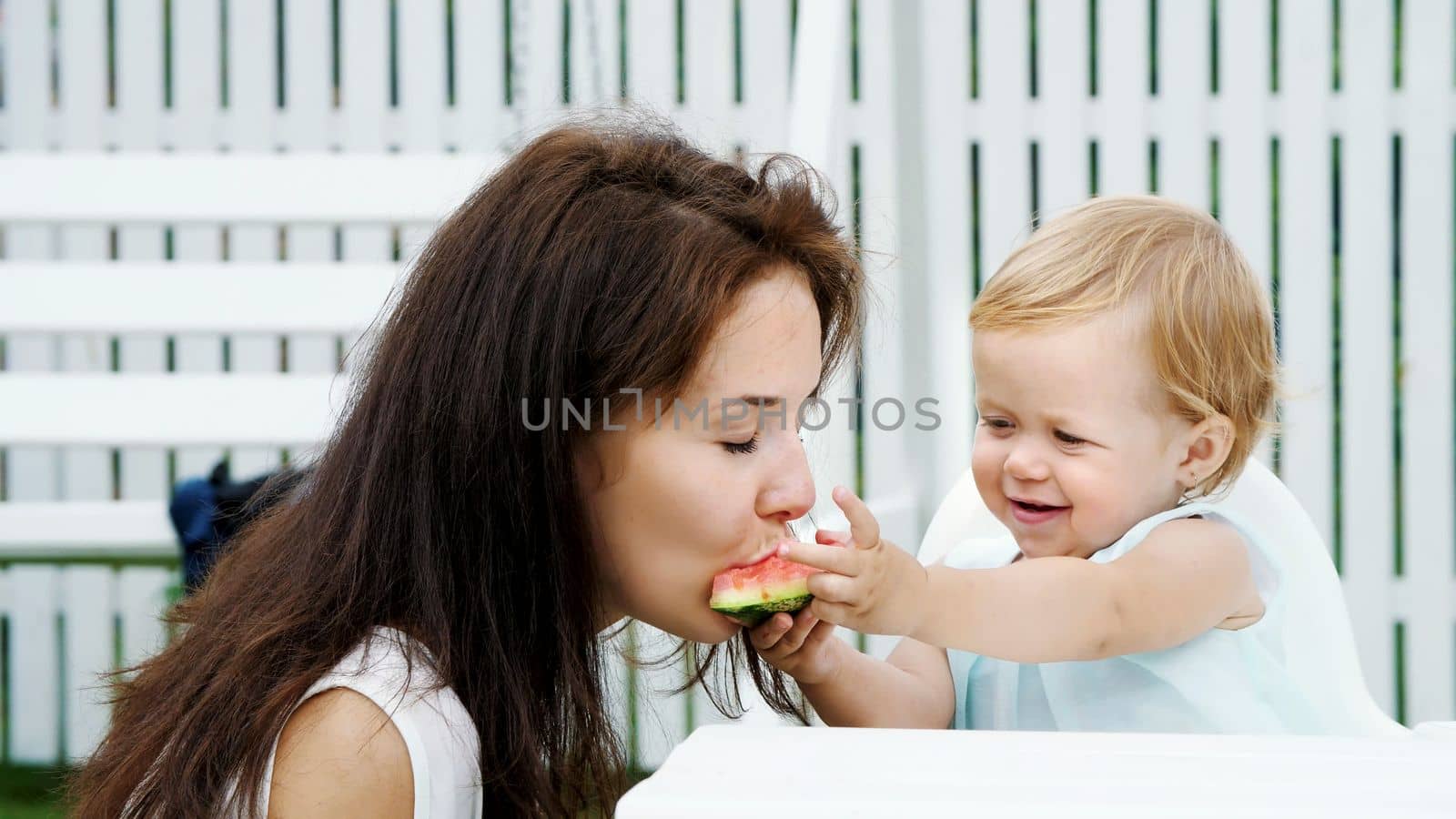 summer, in the garden, a funny one-year-old blonde girl treats her mother with watermelon, feeds her from her hands, the girl also eats watermelon. High quality photo