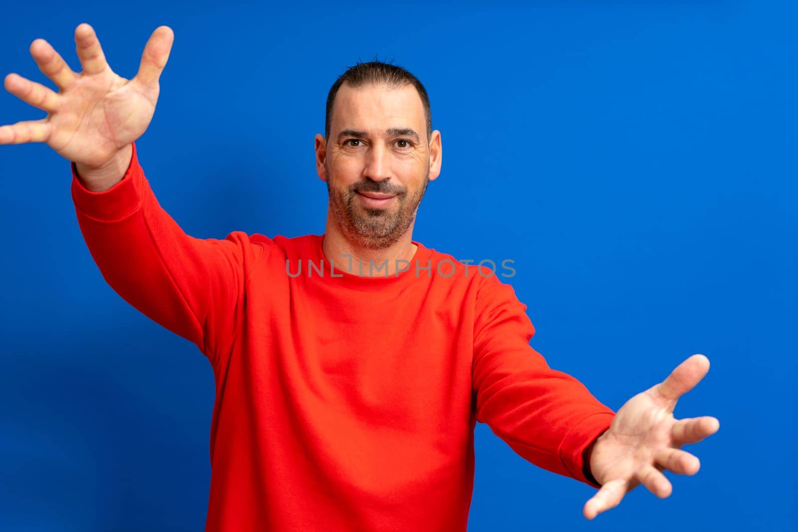 Portrait of a bearded latino man in a red jumper with arms outstretched in welcome isolated over blue studio background