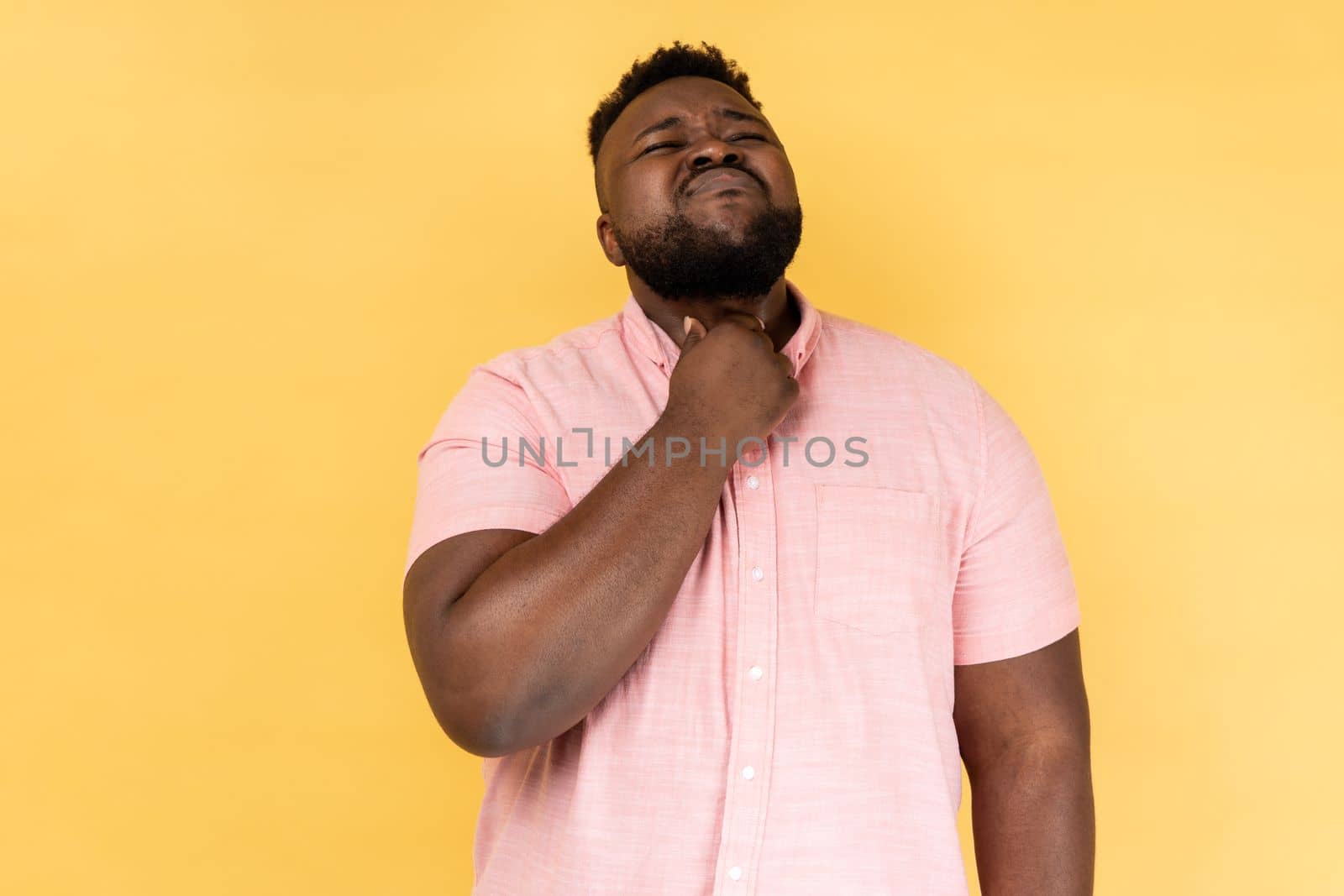 Throat pain. Portrait of sick man wearing pink shirt standing and touching his painful throat, frowning his face, feels terrible pain. Indoor studio shot isolated on yellow background.