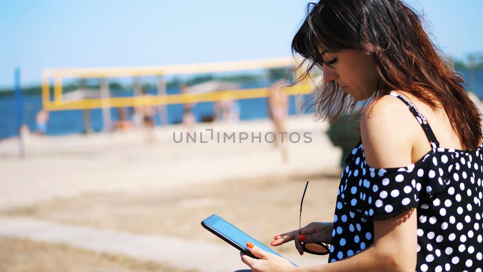 portrait, beautiful girl in sun glasses working on a tablet, on the beach, on a hot summer day, against a background of beach volleyball. High quality photo