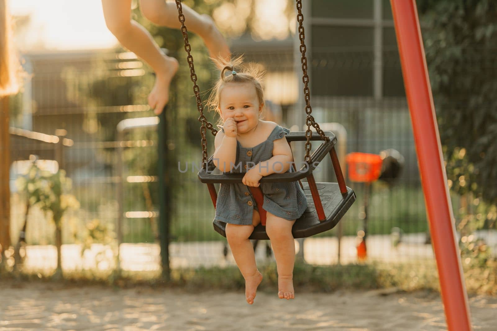 Toddler baby girl on a swing on the warm summer evening. Mother is swinging her young daughter on a sunny playground.