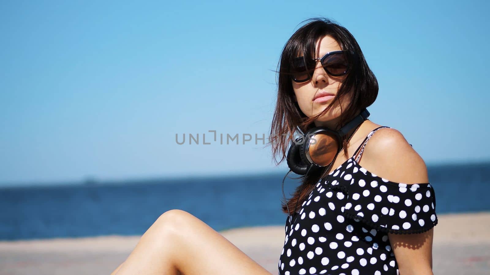 portrait, beautiful brunette, woman in sun glasses, and headphones, sitting on the beach, against the sea. wind waves her hair. High quality photo