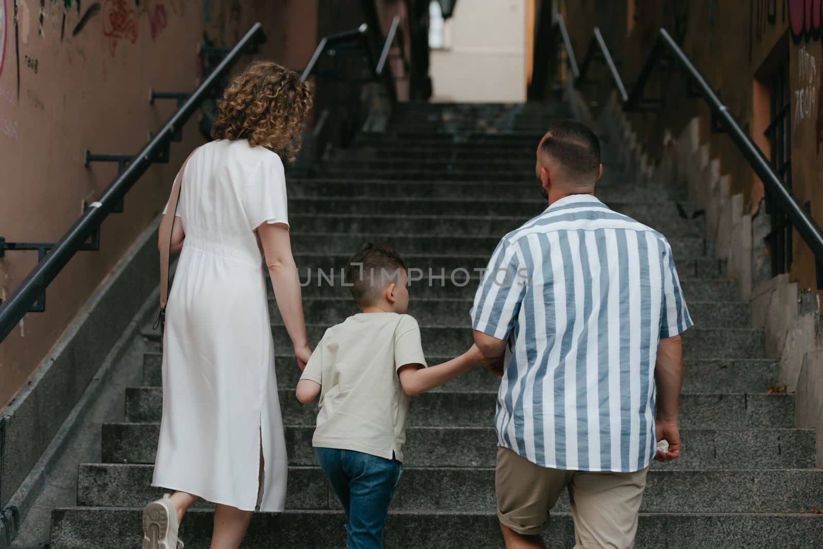 A photo from the back of a family which is climbing stairs in an old European town. A happy father, mother, and son are holding hands and having fun in the evening.