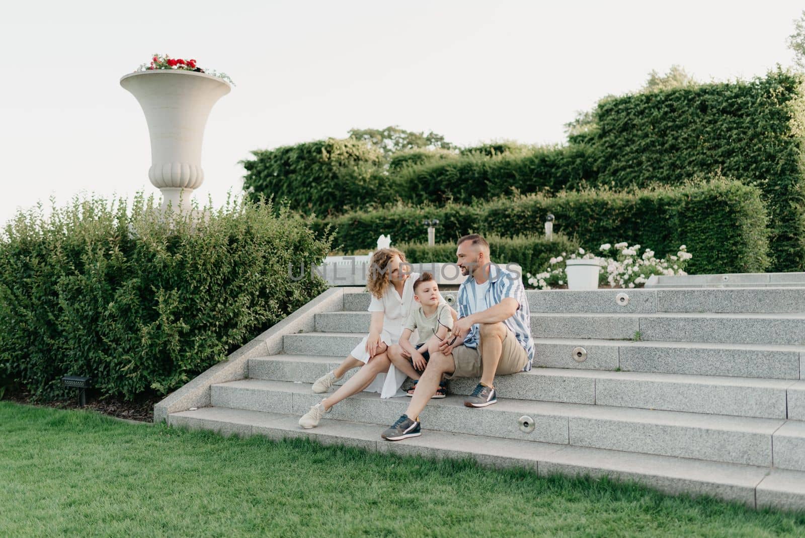 Father, mother, and son are sitting on the steps in the garden of an old European town. Dad is discussing important themes with his smiling family in the park at sunset