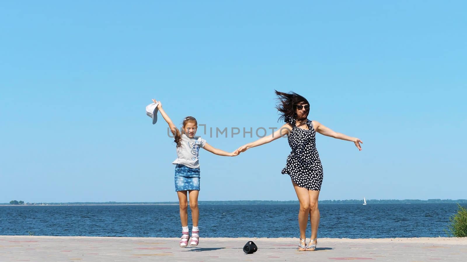 on beach mom and daughter, beautiful brunette in sundress and girl child teenager, dancing, listening to music with mini music bluetooth portable loudspeaker on a hot summer day. High quality photo