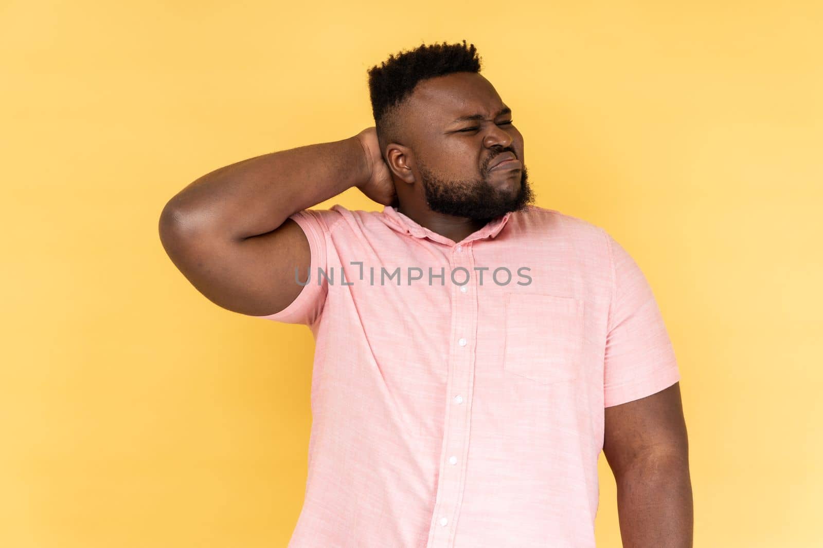 Portrait of worried anxious bearded man wearing pink shirt touching neck, feeling acute pain moving and turning head, suffering spine problems. Indoor studio shot isolated on yellow background.