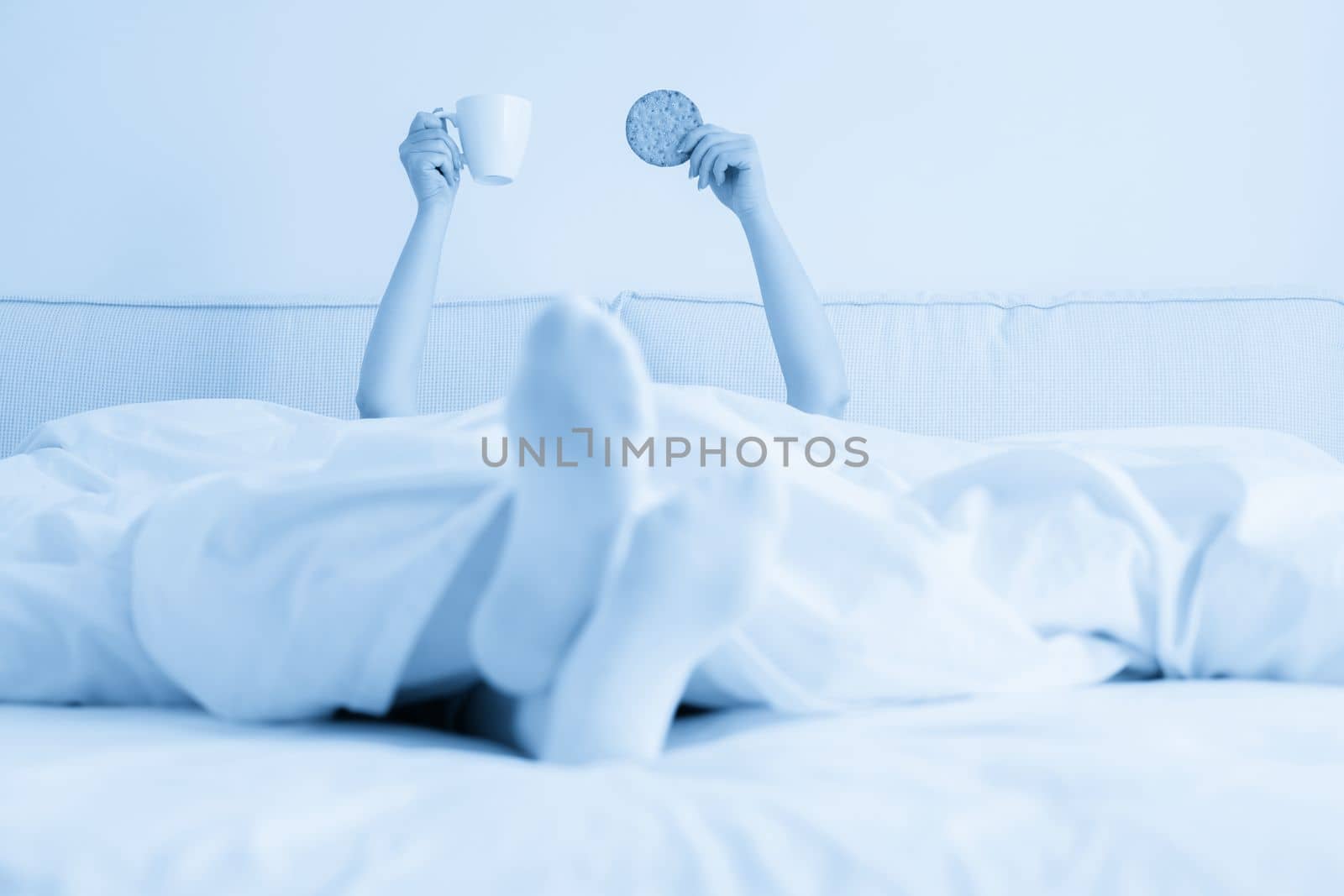Female hand holding cup of coffee from under a blanket in bed. Woman waking up in the morning.
