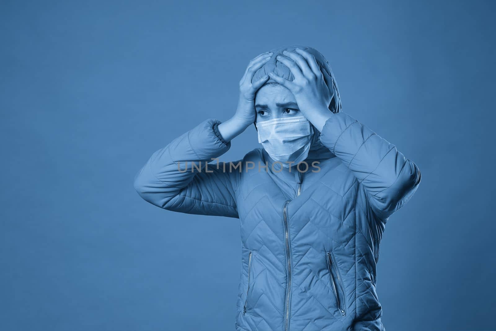 Young brunette woman in protective face mask and coat over blue background. Model girl in medical mask and spring outfit, studio shot with copy space for your text. Anti-flu, anti-epidemic