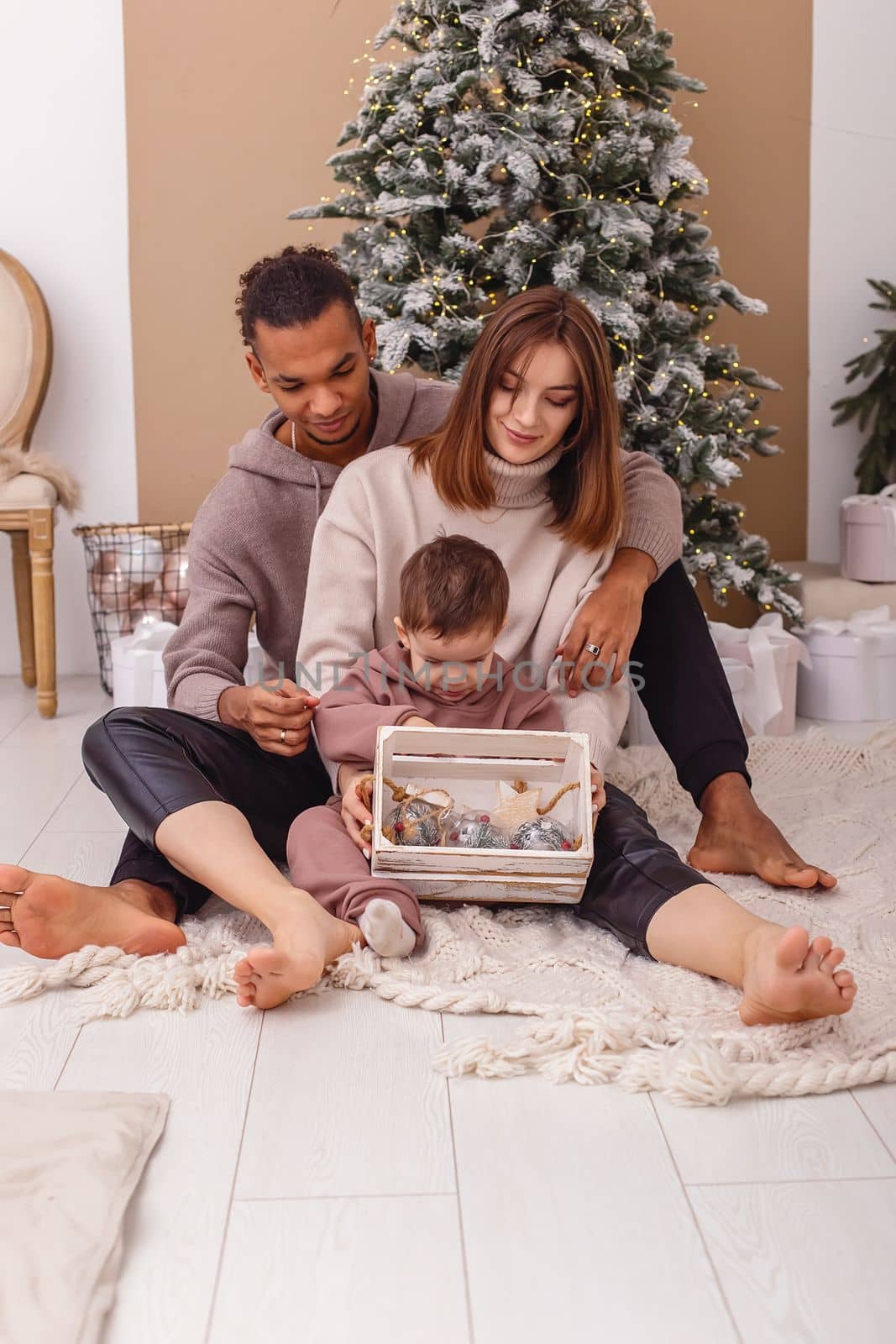 A family is sitting on a knitted blanket, holding a wooden box with Christmas toys. by Zakharova