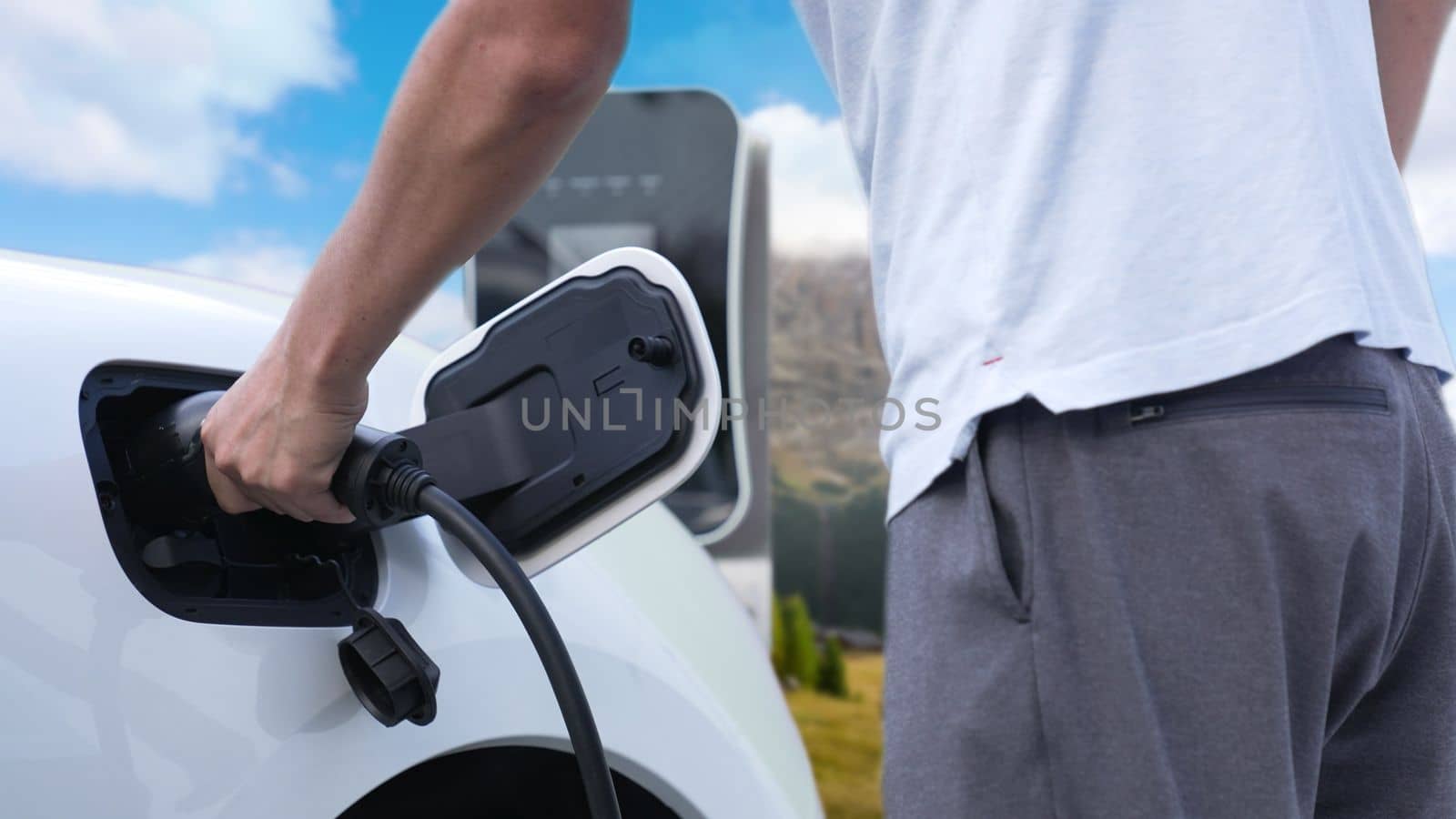 Progressive man recharge electric vehicle installing power cable plug from charging station with rural mountain in the background. EV car driven by electro engine for clean environment3