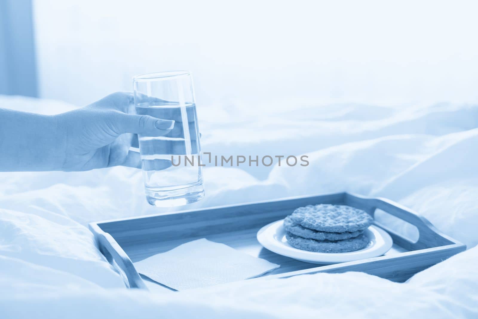 Female hand holding glass of water over tray with crackers on bed by Mariakray