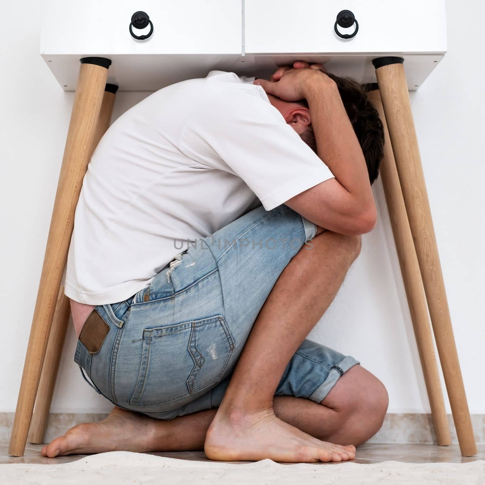 Scared man hiding under table in living room. Live me alone. Stress concept