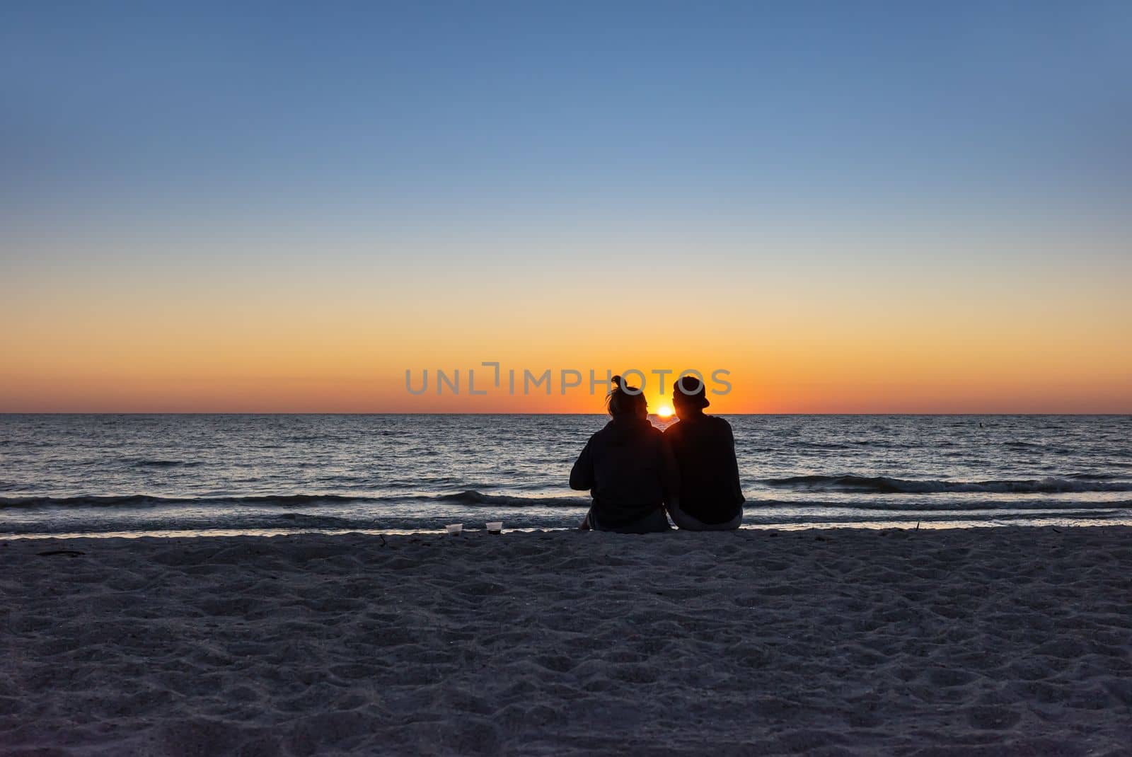St. Petersburg, Florida, USA - Oct. 22, 2022: Beach lovers watching the sunset sitting on a sandy beach.  Beach scene with couple silhouette. Romantic couple on the beach. St. Petersburg, Florida