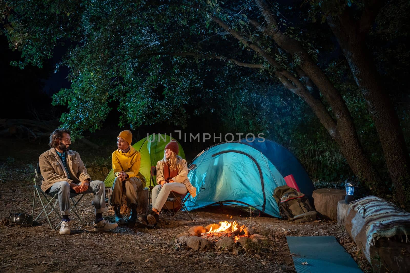 Multiethnic friends sitting around camp fire in the woods. Enjoying nature having fun together camping. Travel and nomad adventure concept. High quality photo