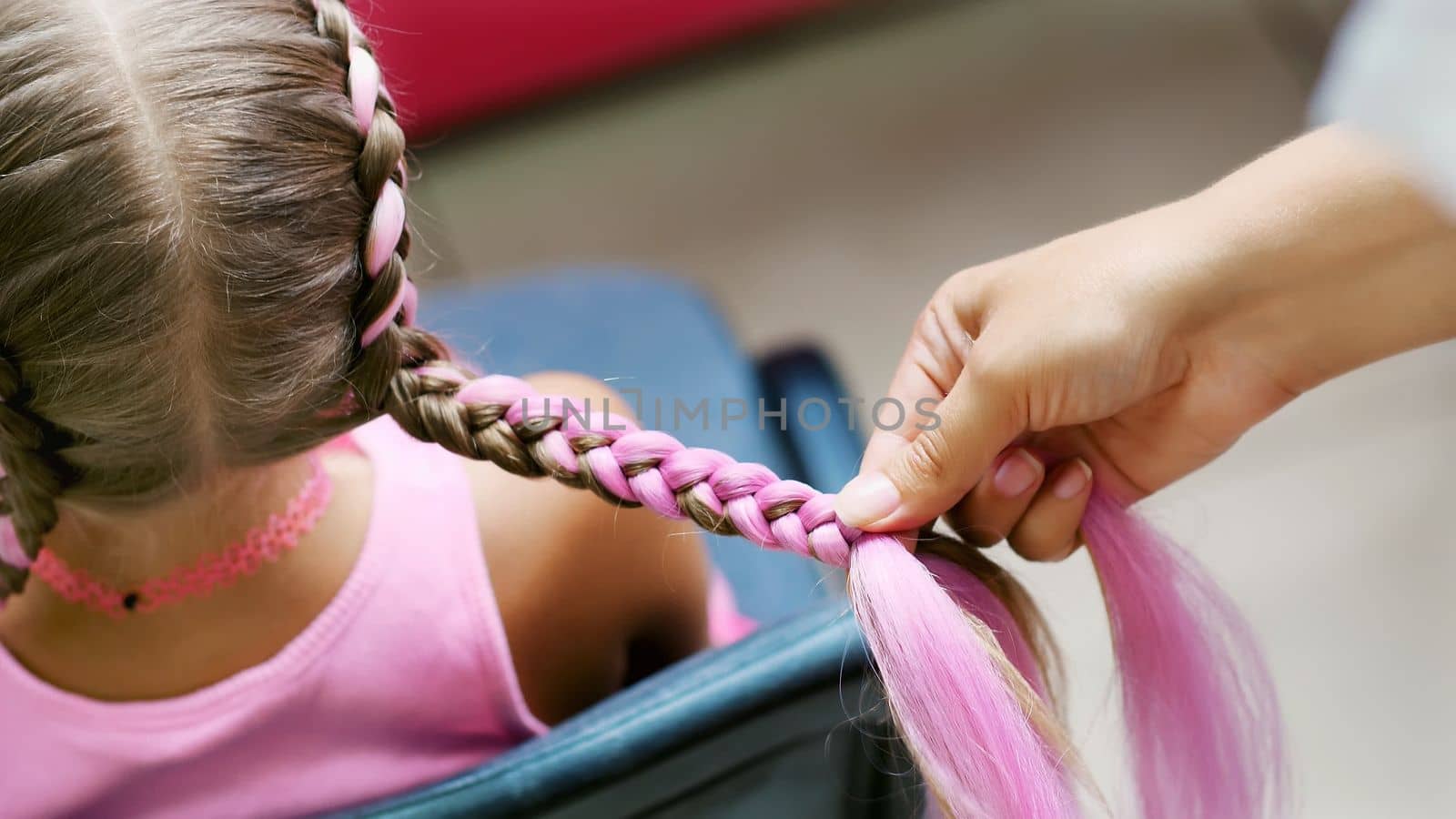 Beautiful blond girl, of seven years old, braided two pigtails, do a hairstyle with pink locks of hair in a beauty salon, a hairdresser's salon, in front of a large mirror. a little princess. High quality photo