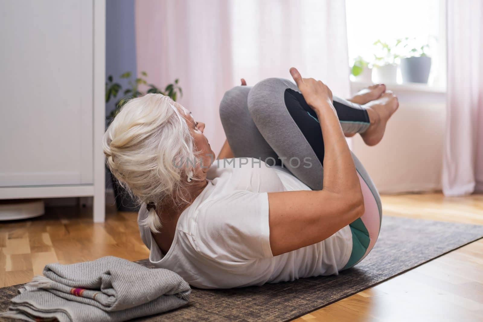 Senior woman lying on yoga mat doing apanasana. Fit female relaxing on floor at home. 