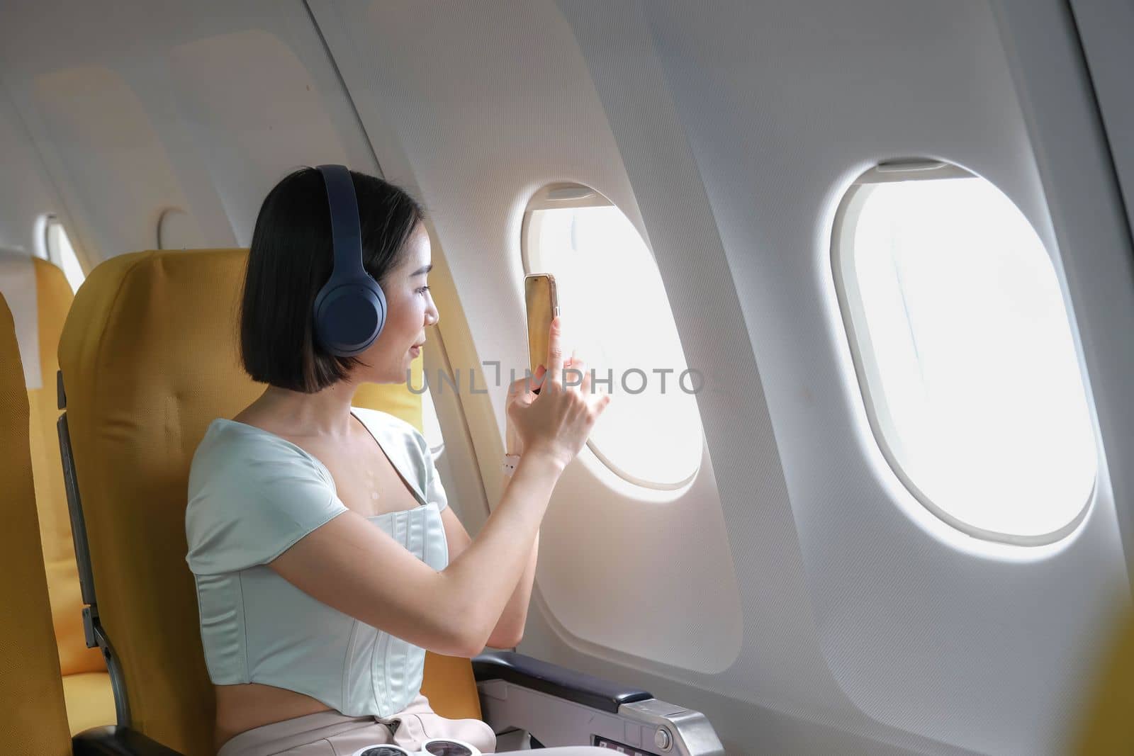 Young female listening favourite songs during flight in first class cabin using mobile playlist and accessory, woman entertaining on airplane board enjoying music in headphones from smartphone..