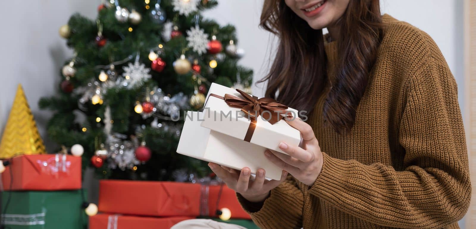 Smiling asian woman in festive mood holding wrapped gift box with brown ribbon in hands, stretching out present to camera, celebrating Merry Christmas